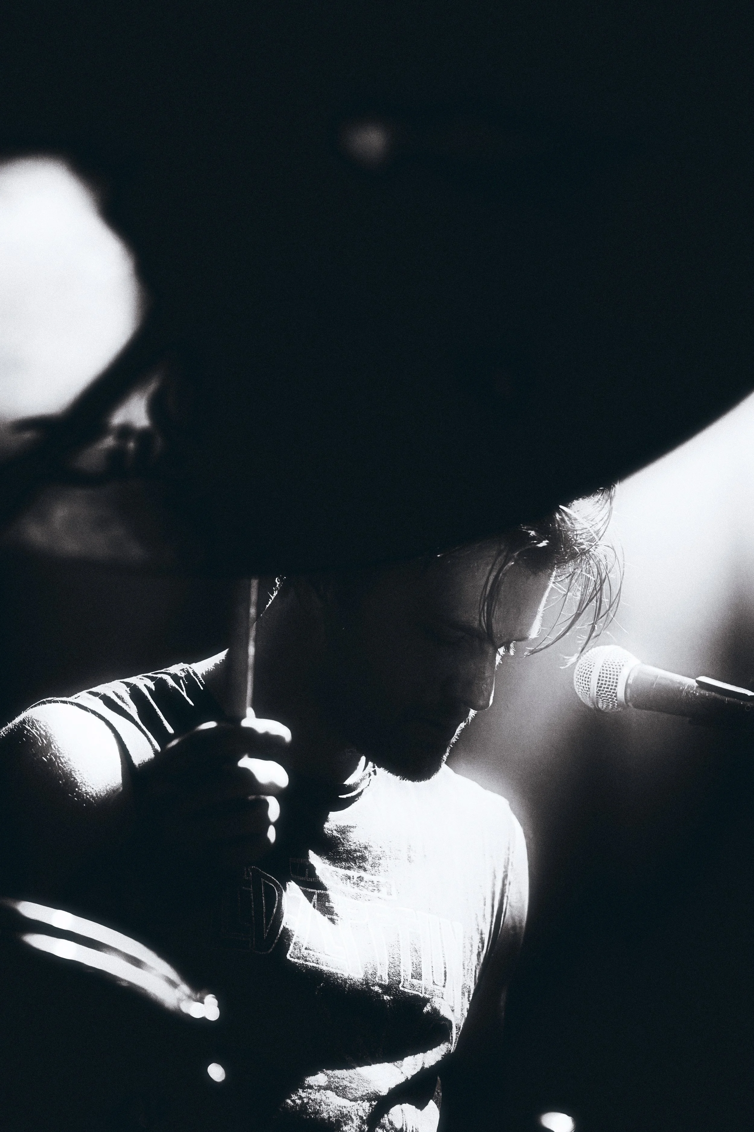 A black and white photo of a person with long hair, holding a microphone close to their mouth, wearing a sleeveless shirt, and standing in front of a microphone stand, with a large hat or object partially obscuring their face.