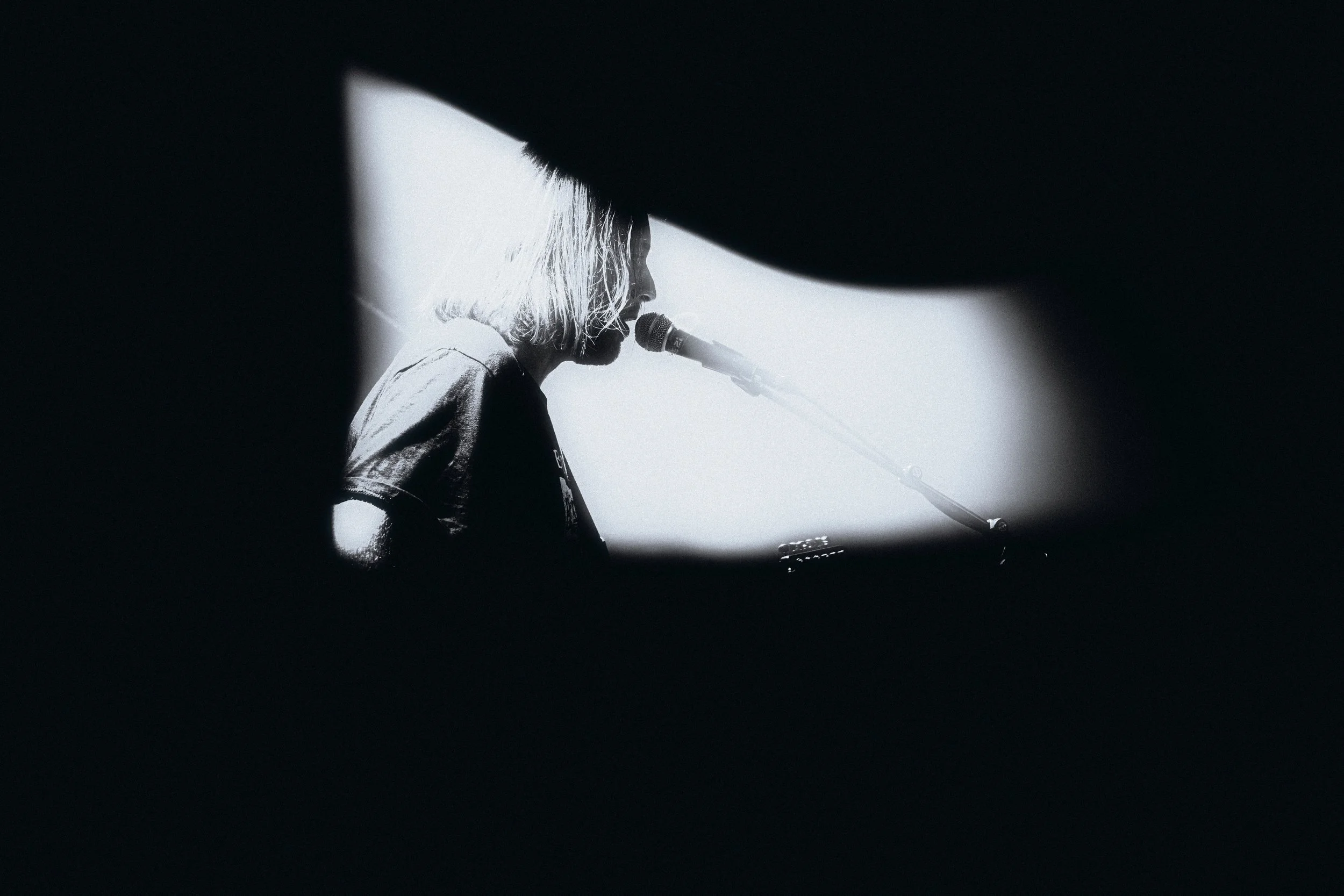 Black and white photo of a person with shoulder-length hair singing into a microphone, viewed through an opening in a dark object.