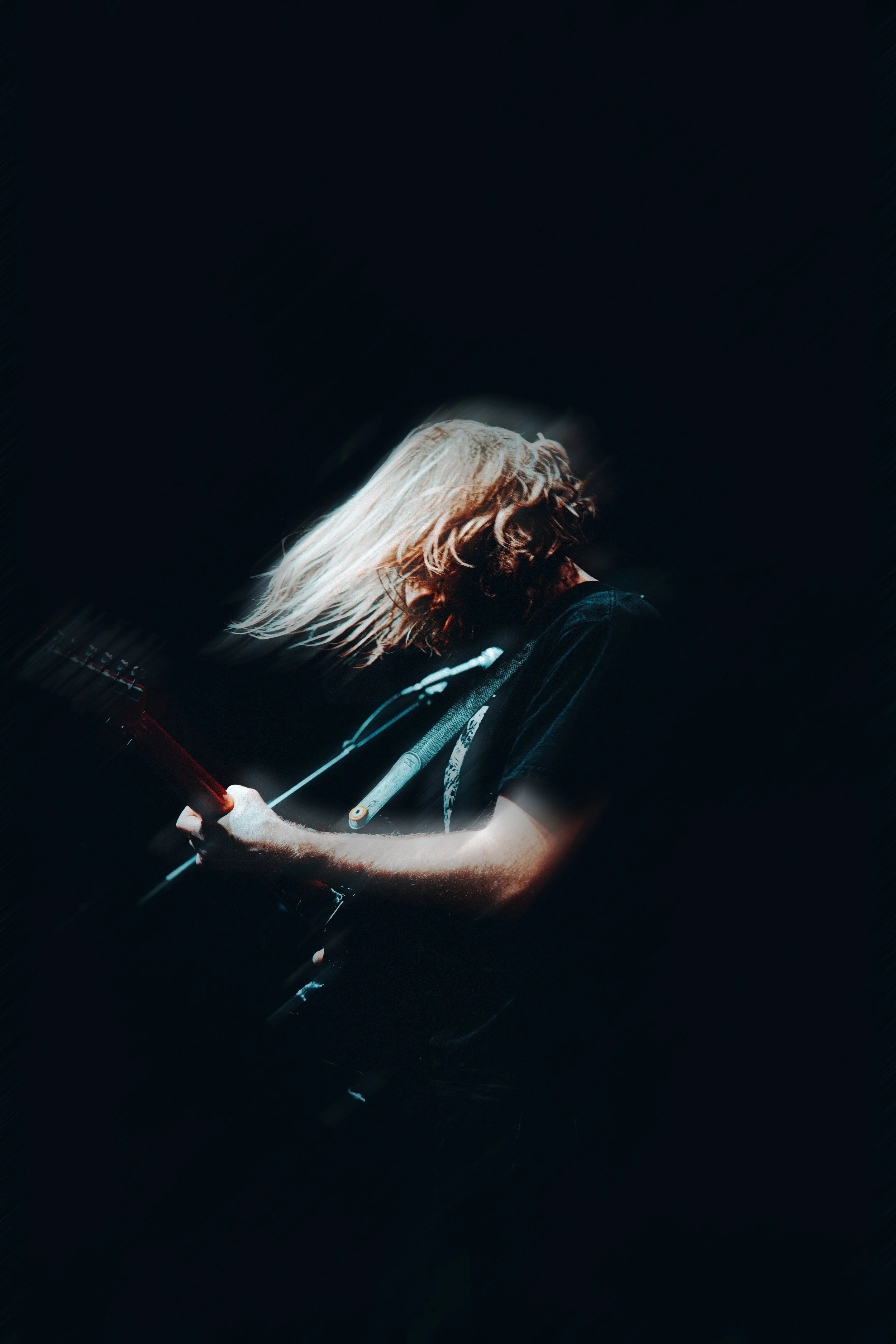 Person with blonde and brown hair playing an electric guitar, head tilted downward, dark background.