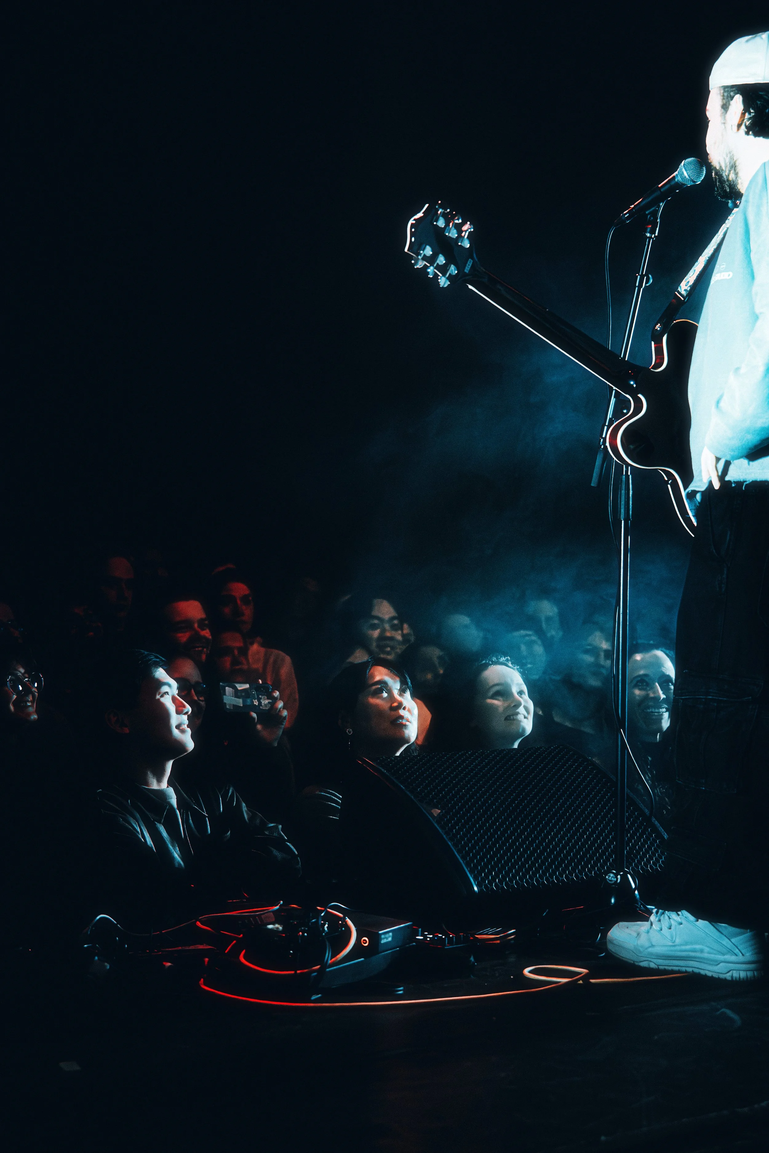 A performer with a guitar on stage facing an audience of diverse people who are smiling and looking at the performer, illuminated by stage lighting.