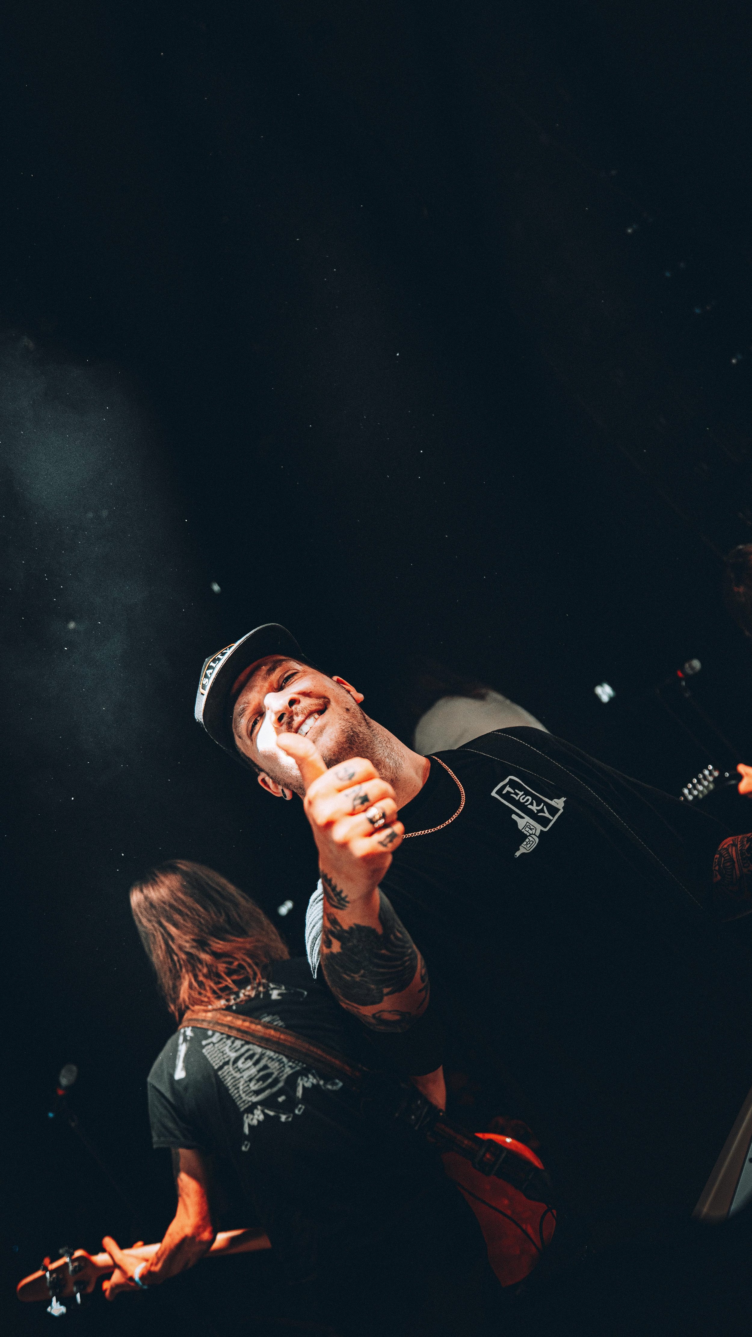 A man smiling and giving a thumbs up at a concert or event, with a person playing guitar in the background, under dark lighting.