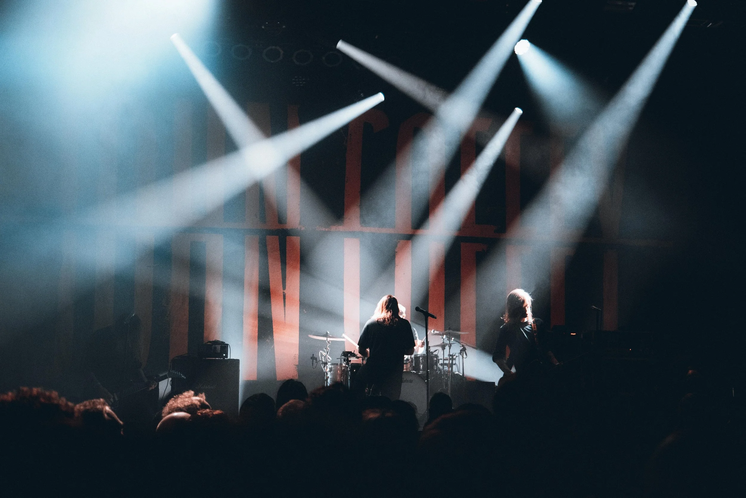 A live concert performance on stage with bright lights shining down on musicians playing instruments, with an audience in the foreground.