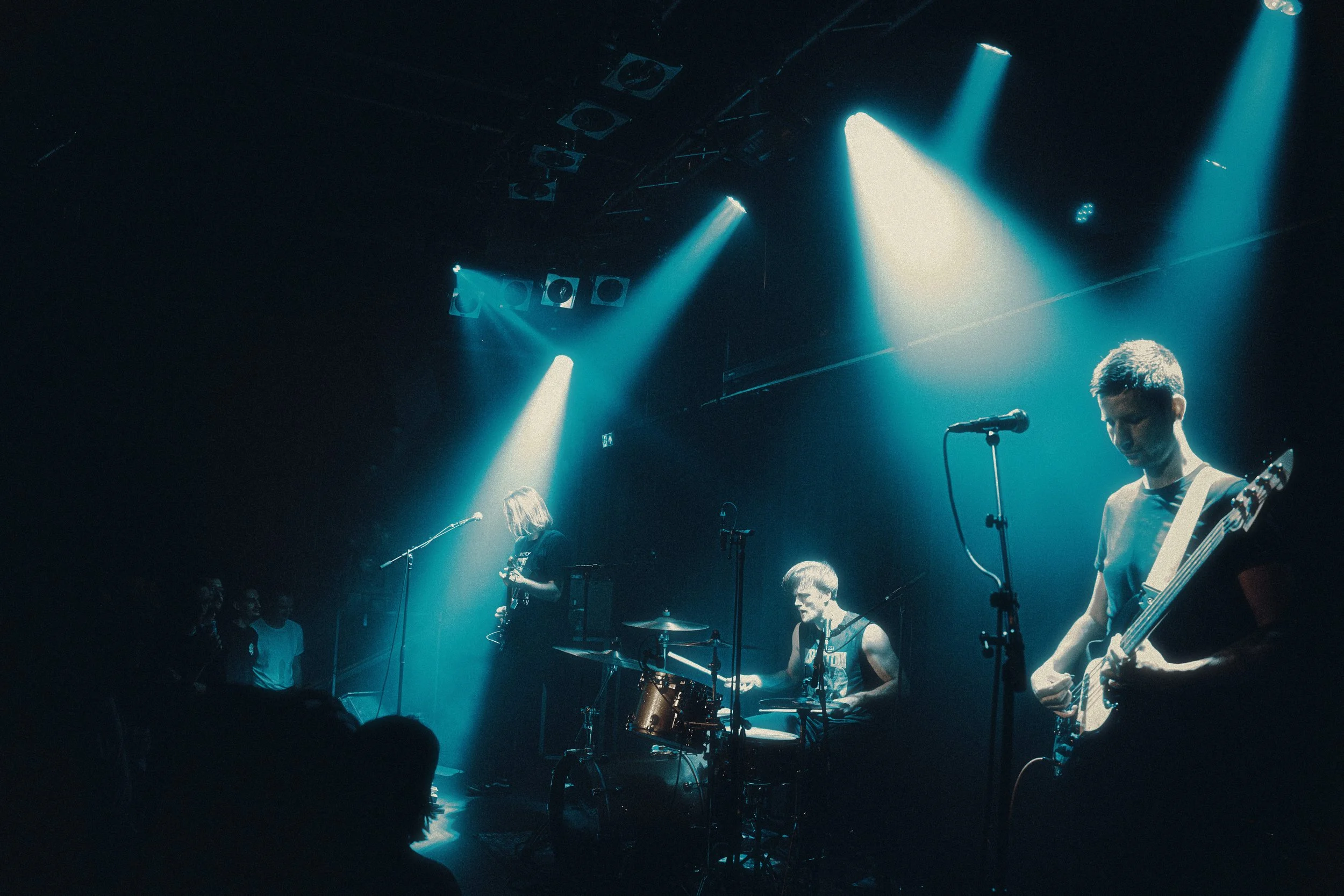 A band performing on stage with blue and white lighting, featuring a drummer, guitarist, and vocalist, with an audience watching in the dark.