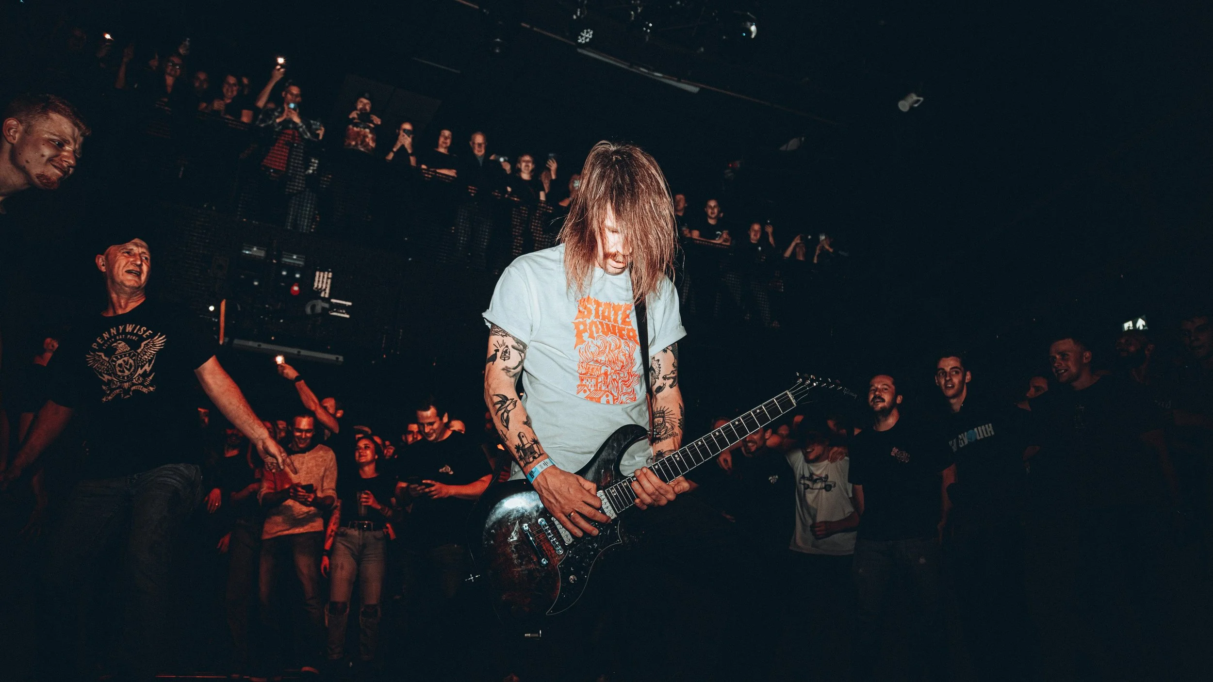A musician with long hair and tattoos playing an electric guitar on stage at a concert, surrounded by a cheering crowd in a dark venue.