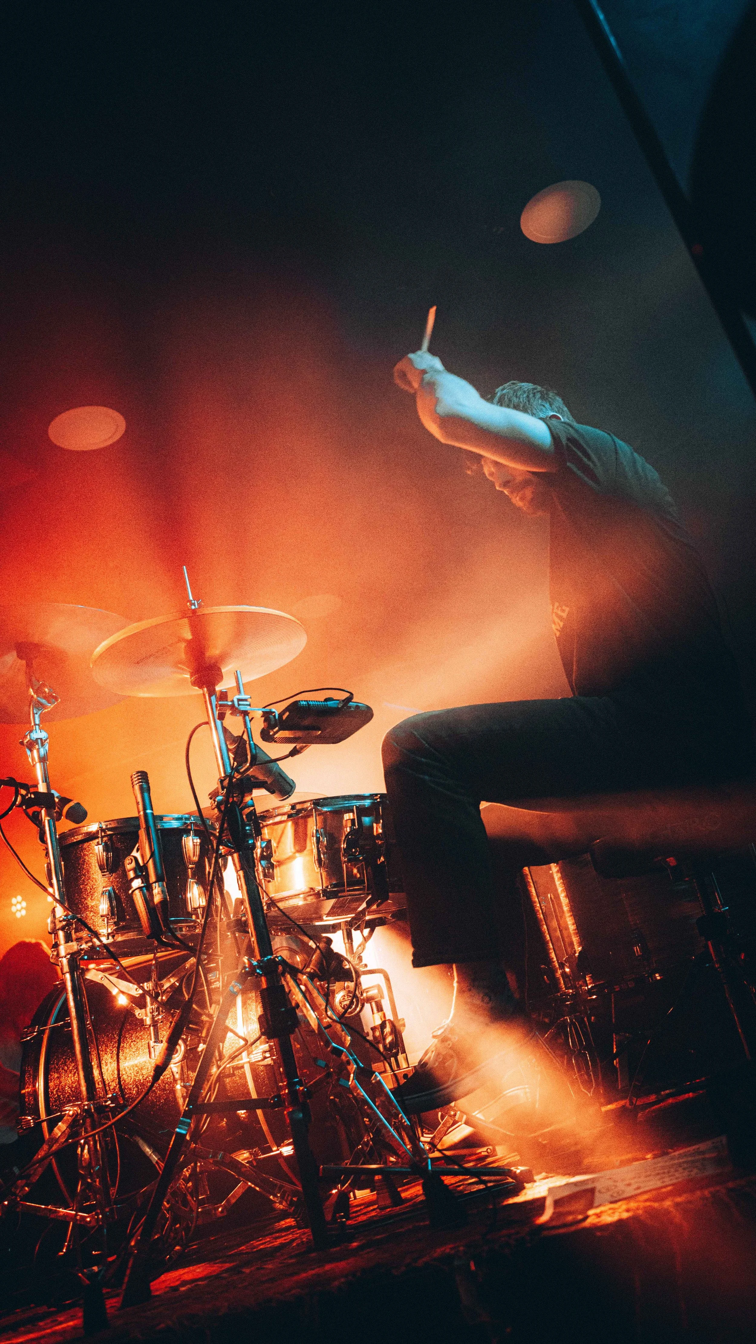 A drummer playing a drum kit on stage with orange and blue lighting effects.