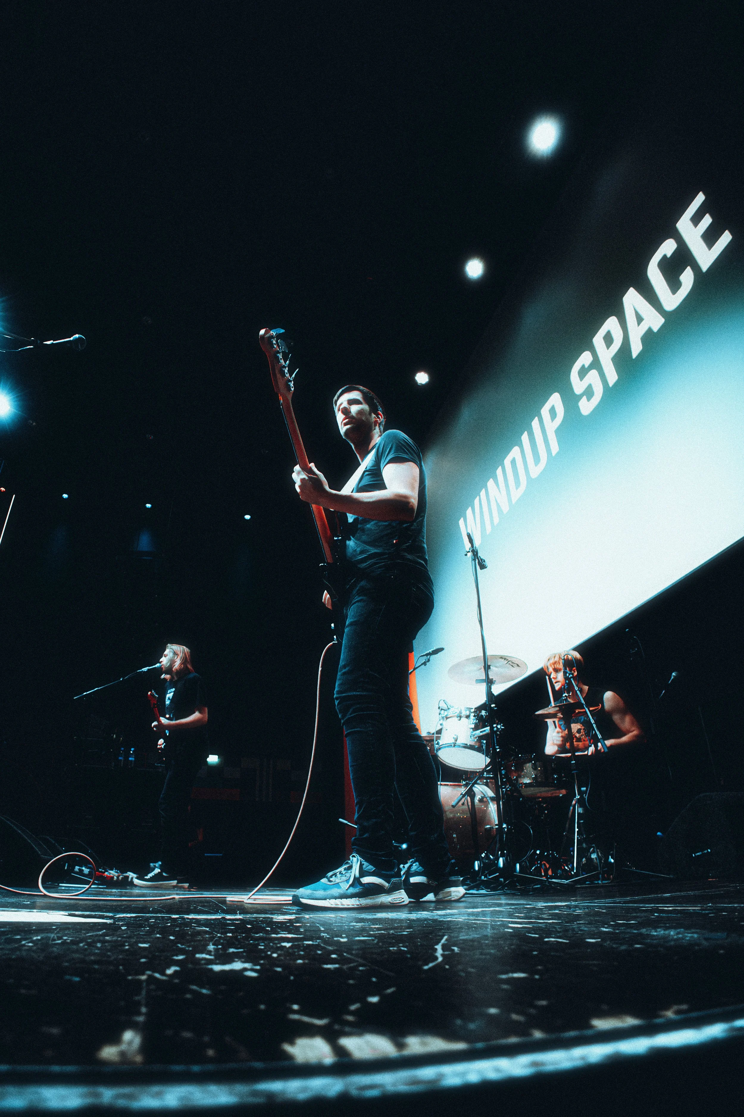 Band performing on stage with a large screen in the background displaying 'WINDUP SPACE'