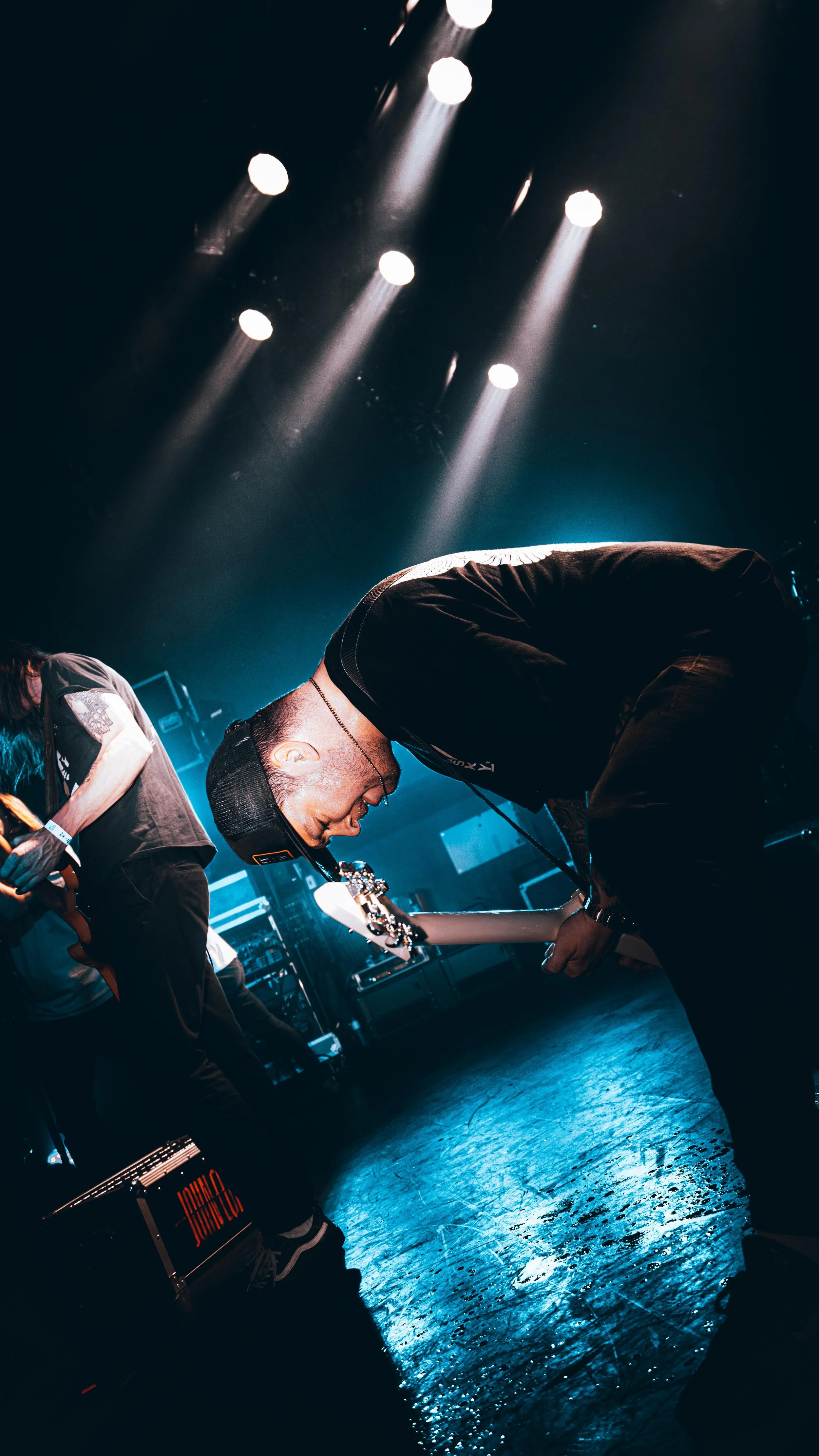 A male performer bending forward on stage, illuminated by blue stage lights, with other band members and equipment in the background.