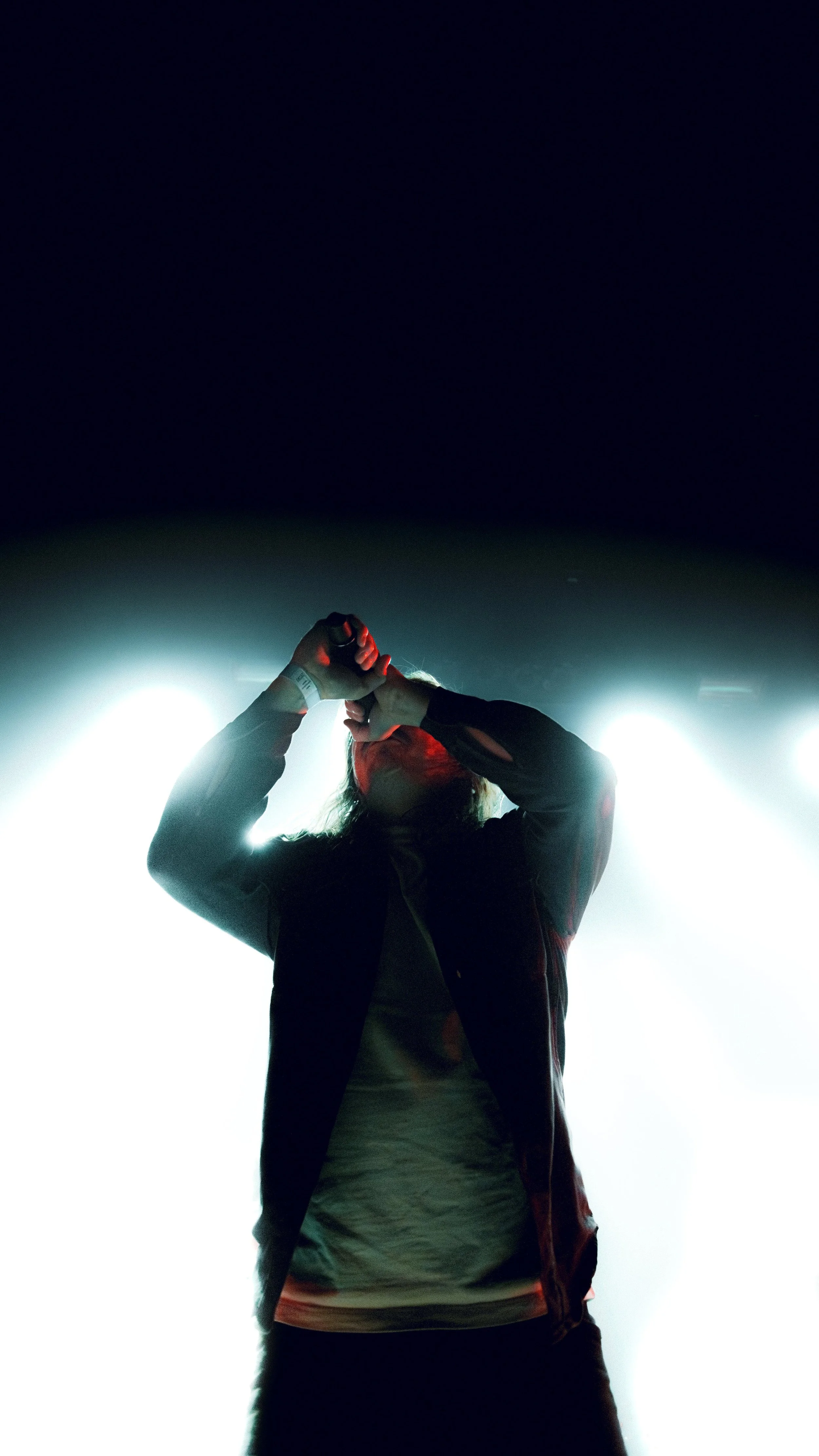 Person standing on stage with bright backlighting, facing downward and adjusting their wristwatch.