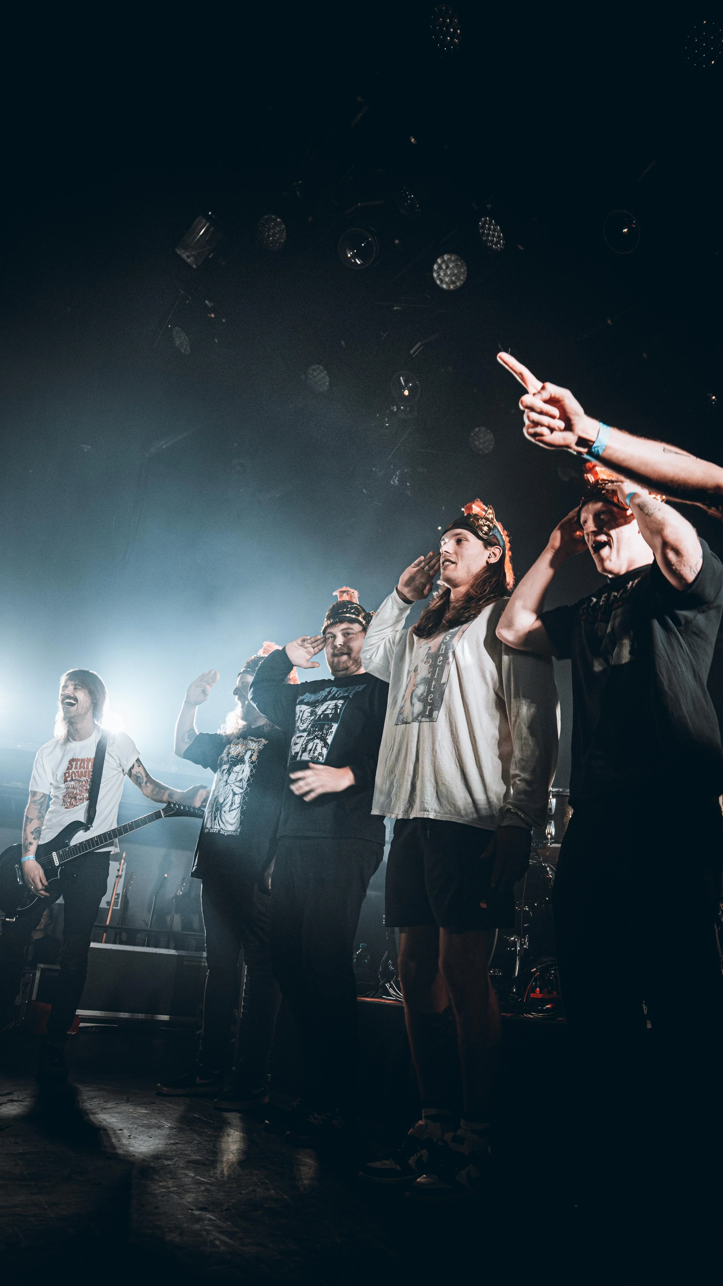 Group of people on stage at a concert, some saluting and others raising their hands, with bright stage lights overhead.