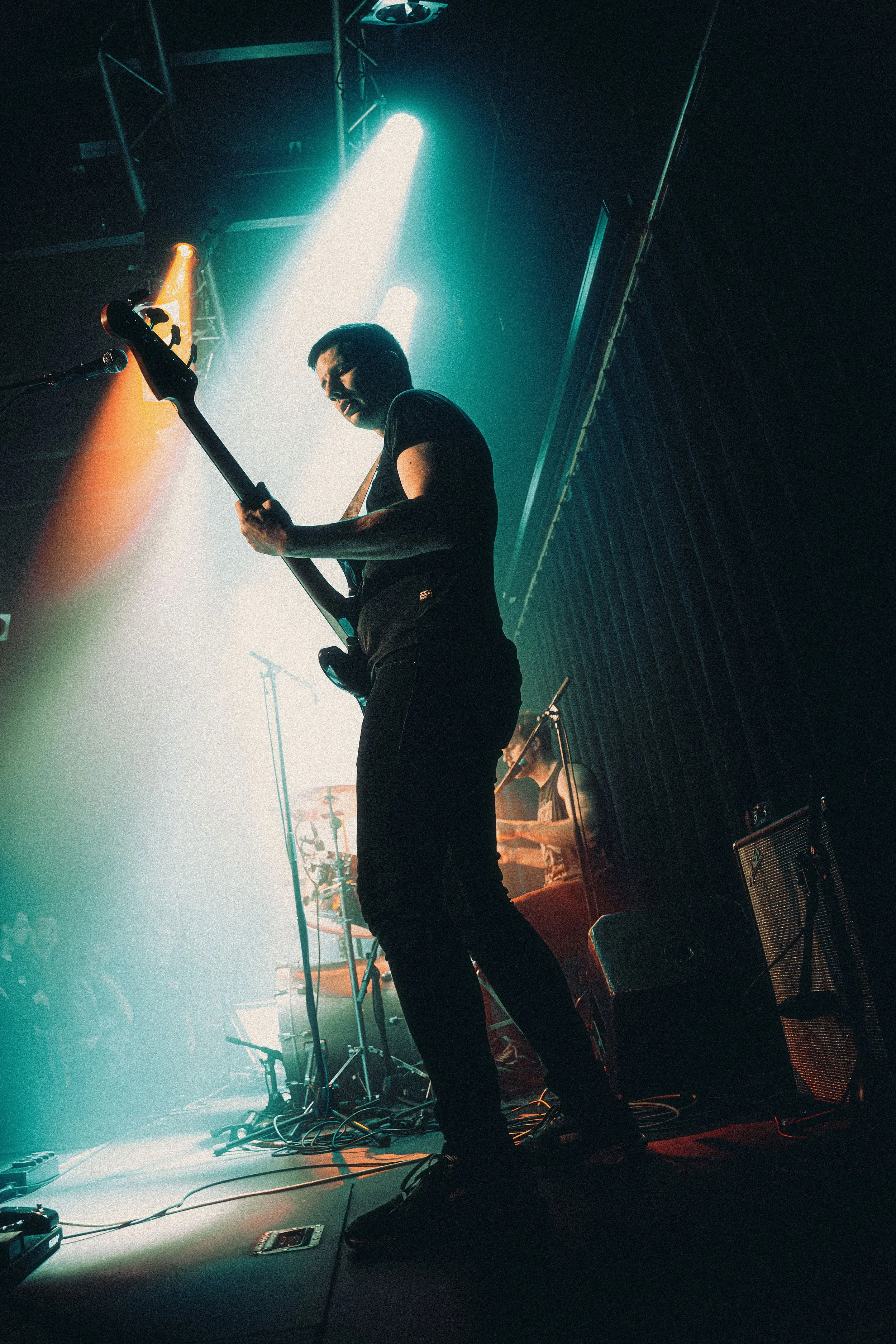 A musician playing an electric guitar on stage, with colorful stage lights shining down, and a blurred drummer in the background, during a live performance.