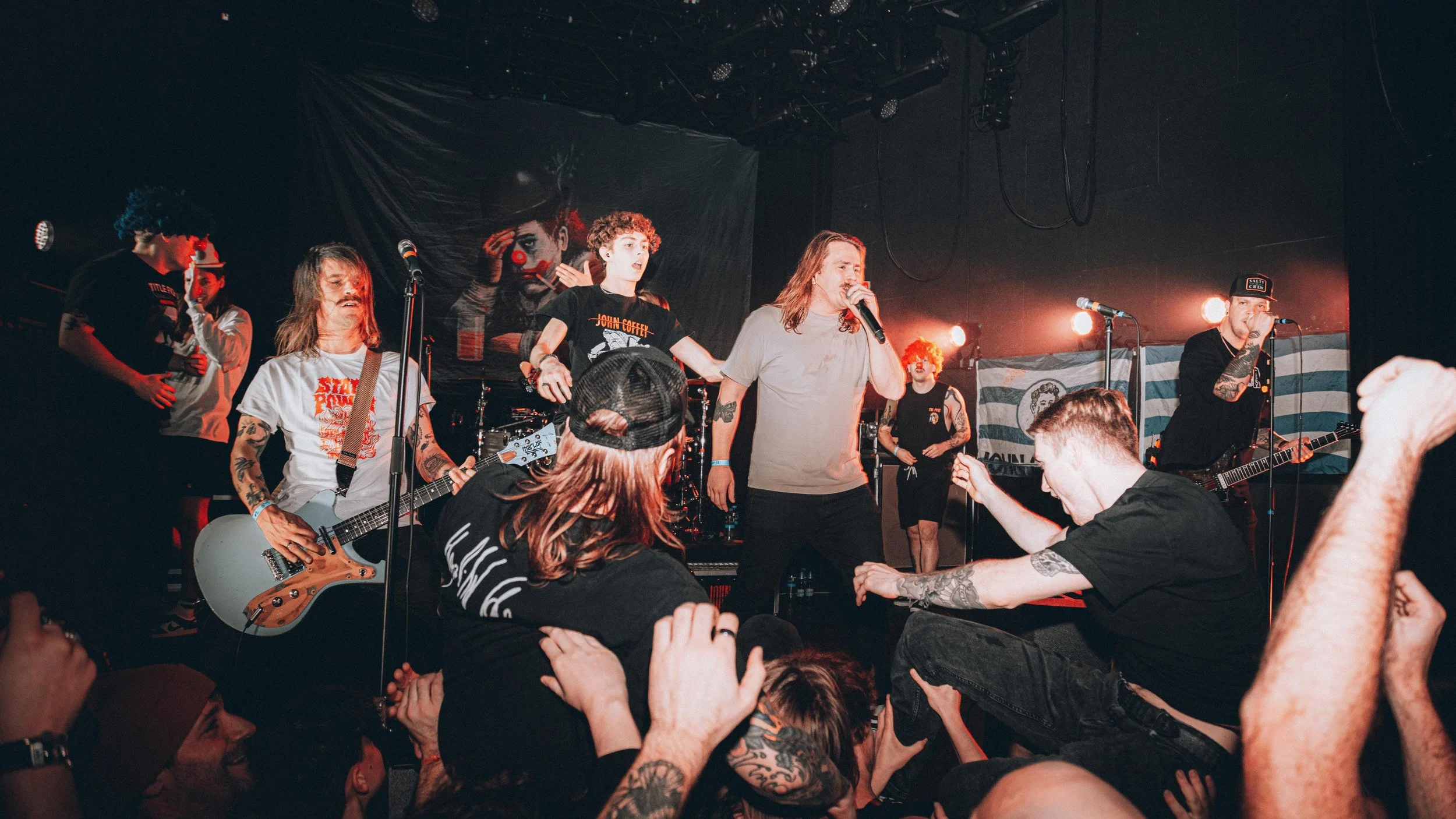 A live music concert with a band performing on stage, including guitarists, vocalists, and a drummer, in a dimly lit venue with audience members close to the stage. The backdrop features artwork of a clown and a flag with stripes.