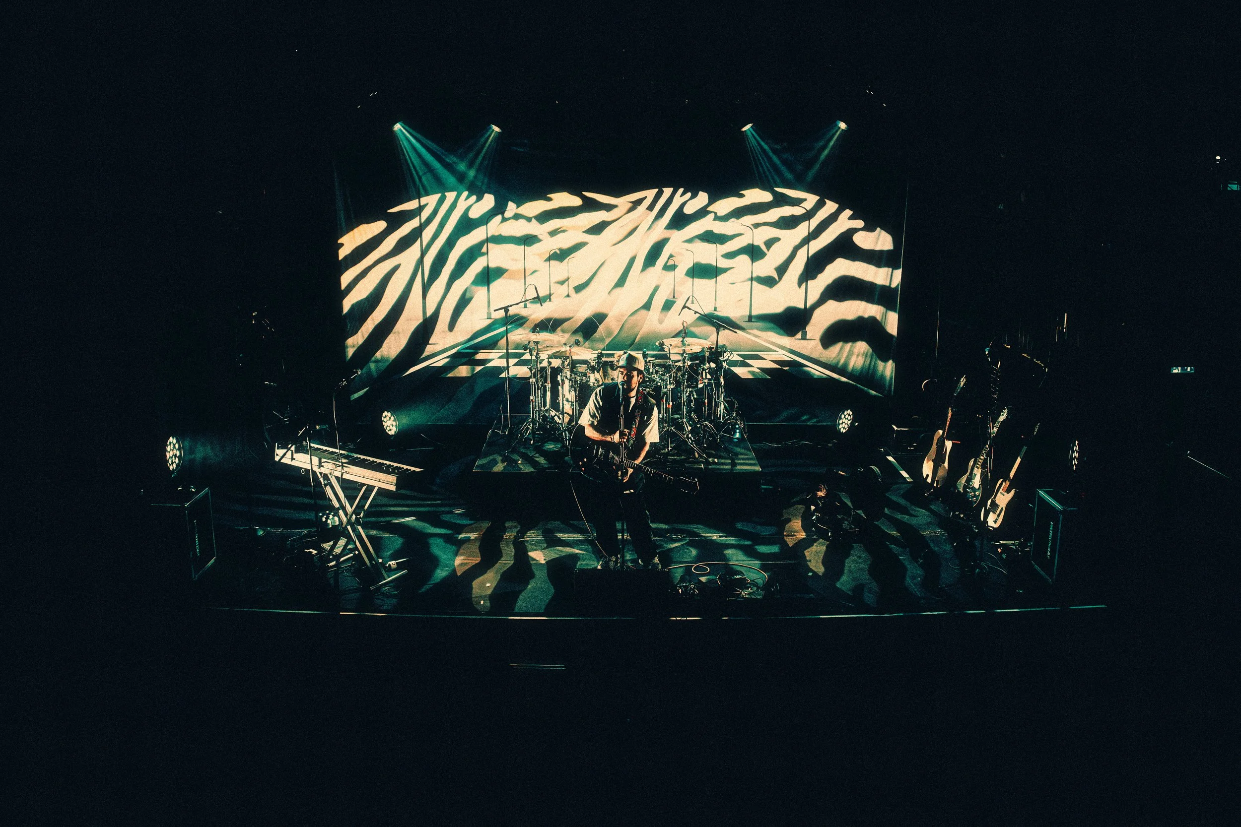 A musician performing on stage with a guitar, surrounded by musical instruments including drums, keyboards, and multiple guitars, with stage lighting and a large screen displaying zebra stripe patterns in the background.