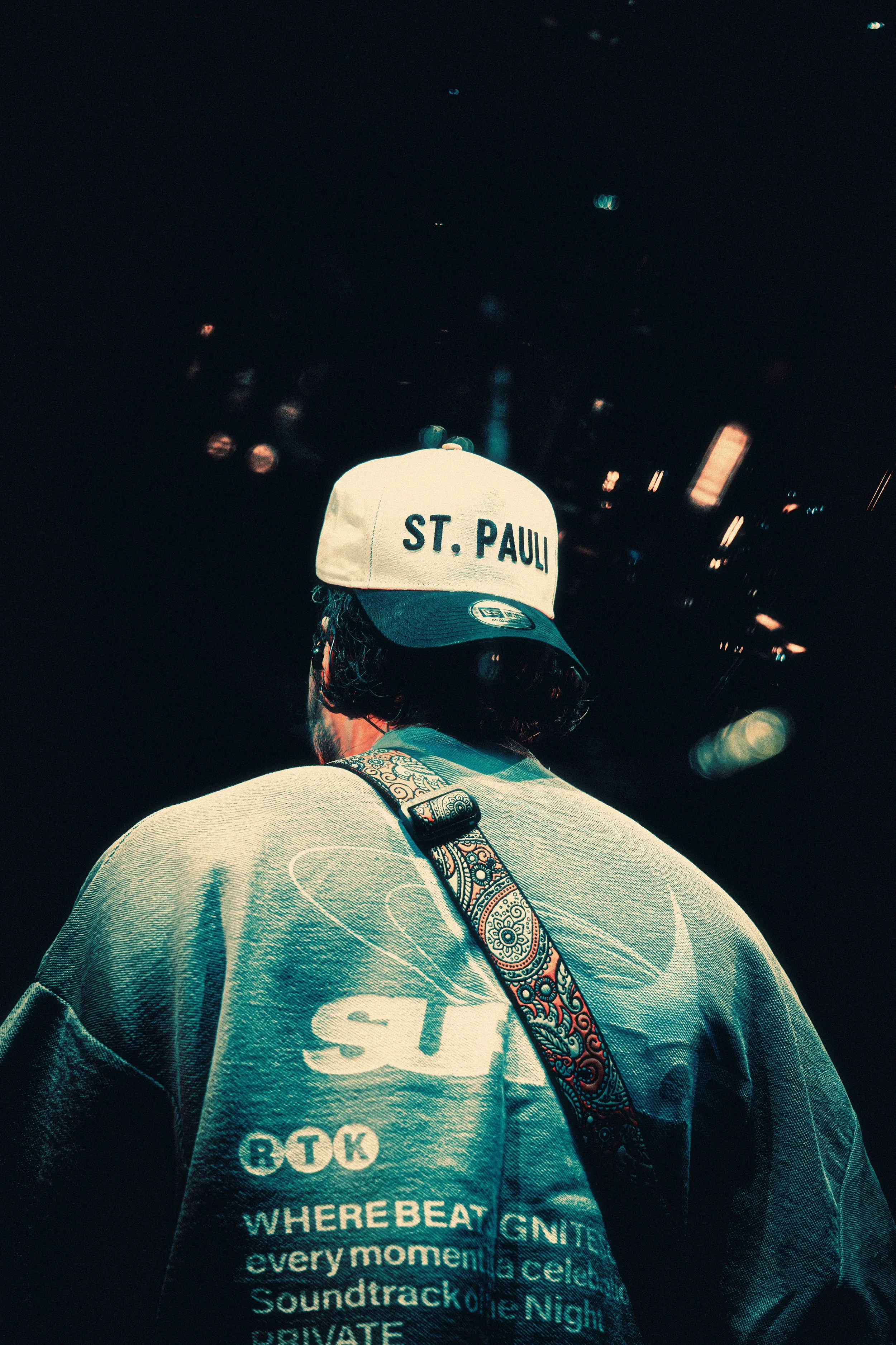 Back of a person wearing a St. Pauli cap, a grey sweatshirt with various logos and text, and a patterned strap across their back, against a city nightscape with blurred lights.