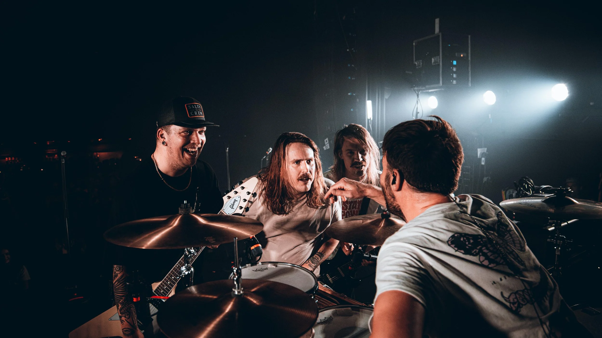 Four musicians on stage in a dark venue, with one playing drums and the others holding guitars, engaged in a heated discussion or confrontation.