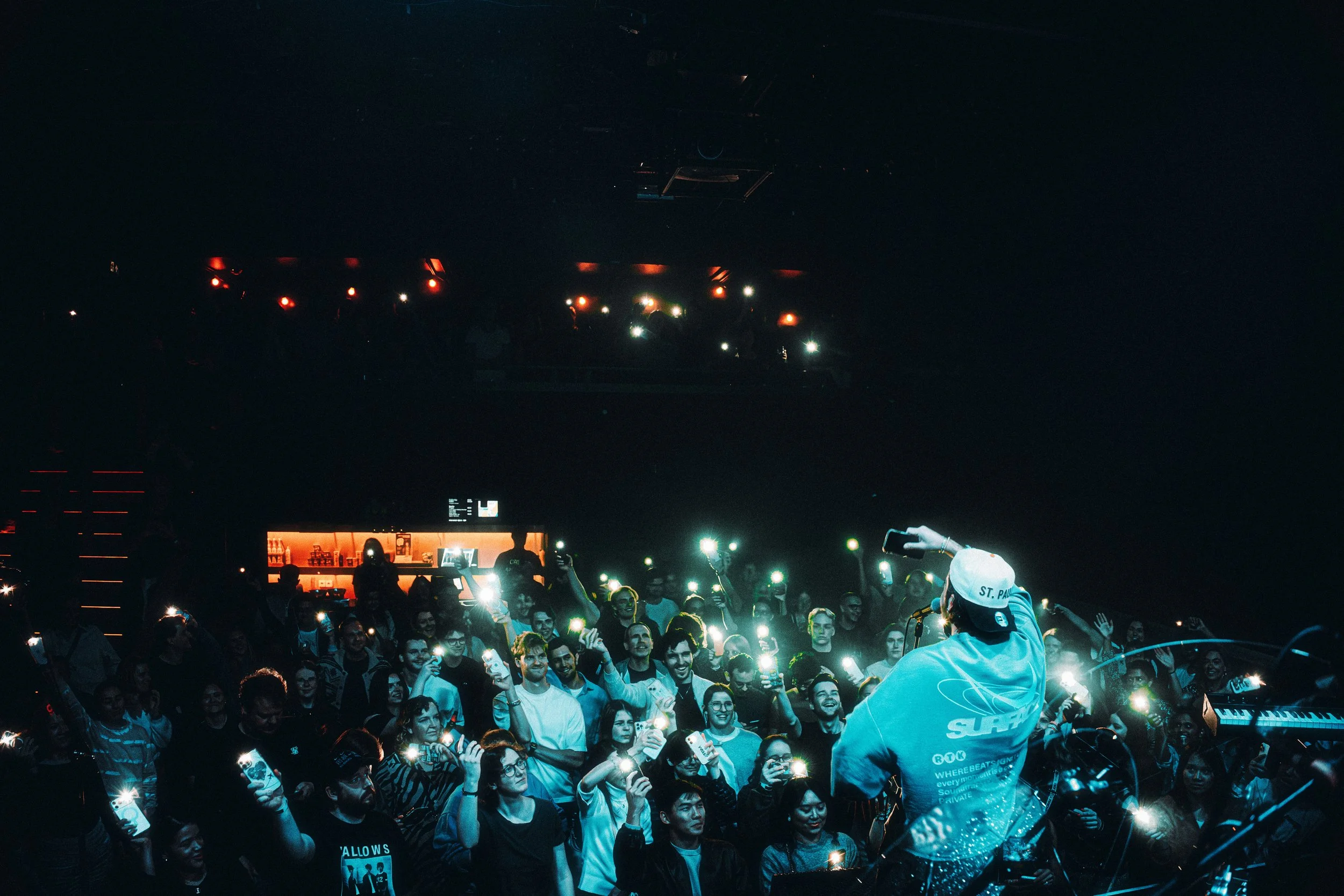 A DJ performs on stage at a nightclub or concert venue, facing a crowd of people holding up smartphones with flashlights on. The crowd is smiling and enjoying the music in a darkened environment with some stage lights.