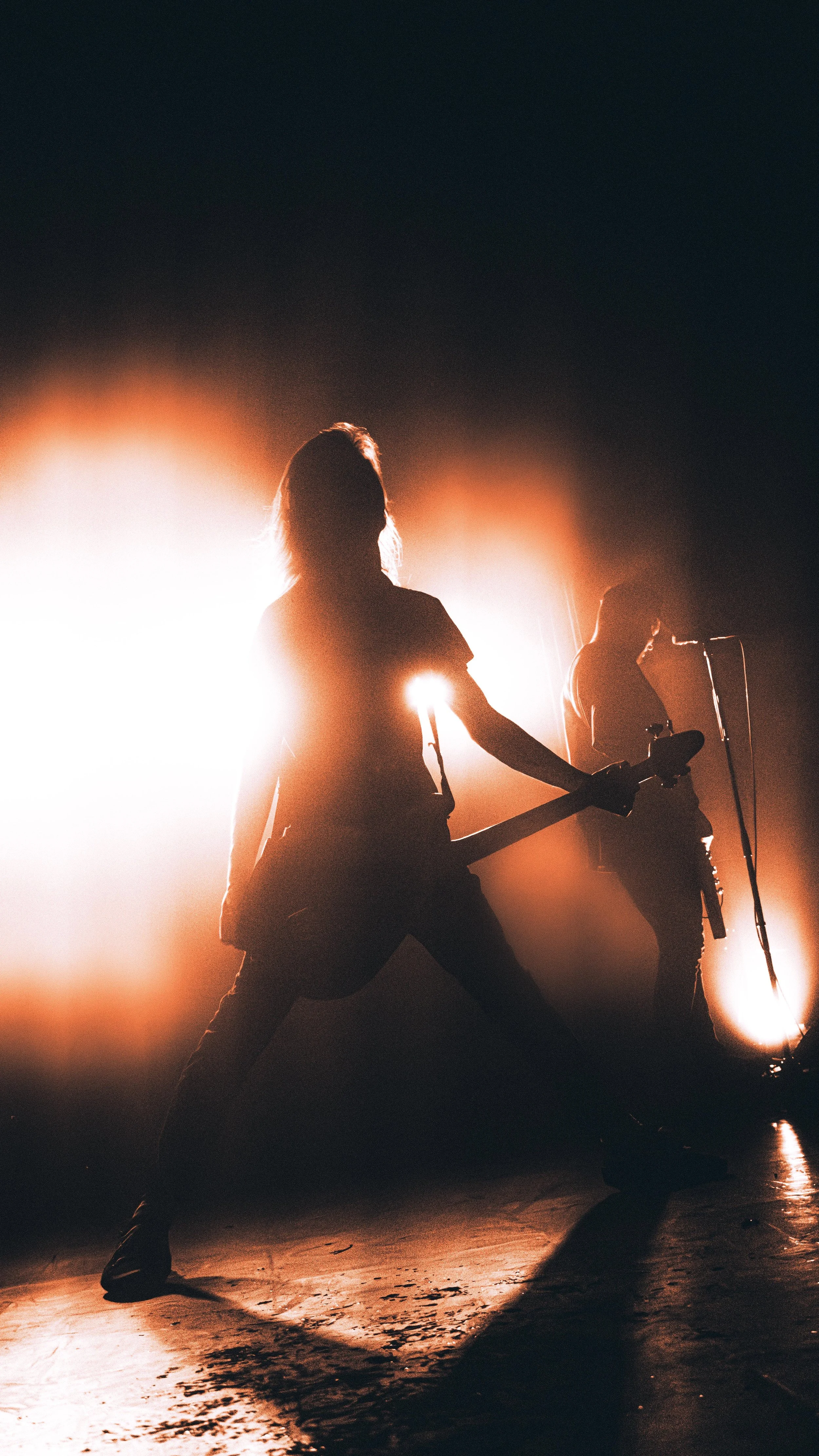 Silhouettes of a female guitarist and a male singer performing on stage with bright backlighting, creating a dramatic scene.