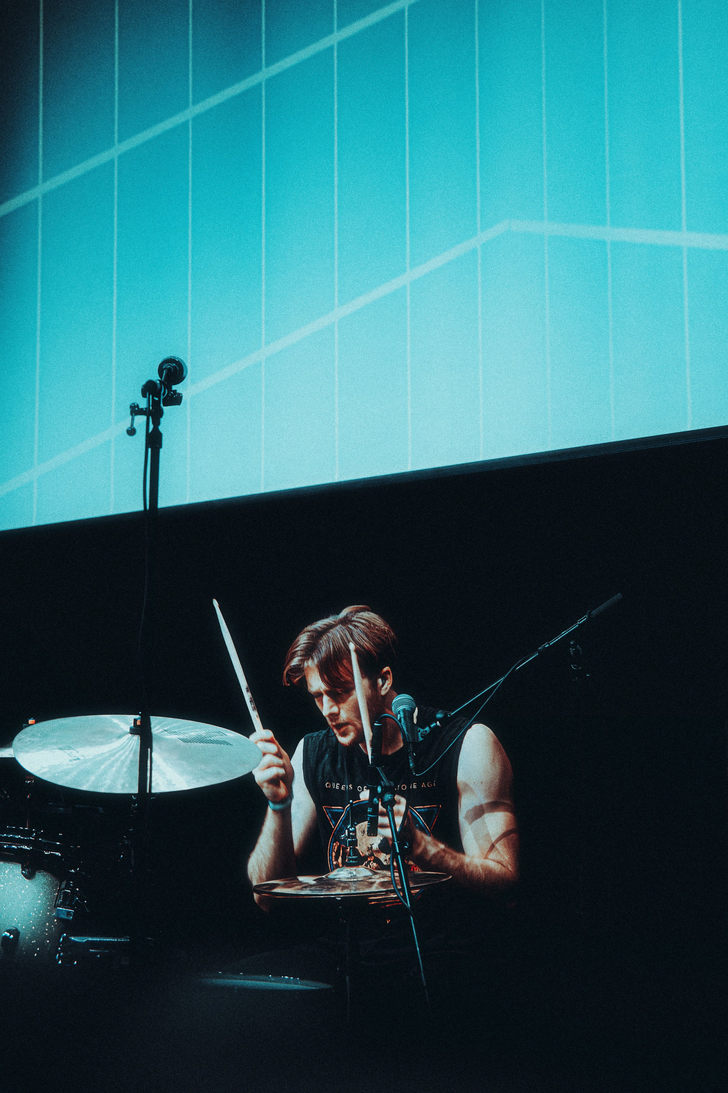 A young man playing drums on stage with a microphone, wearing a black sleeveless shirt, with a large screen displaying a graph behind him.