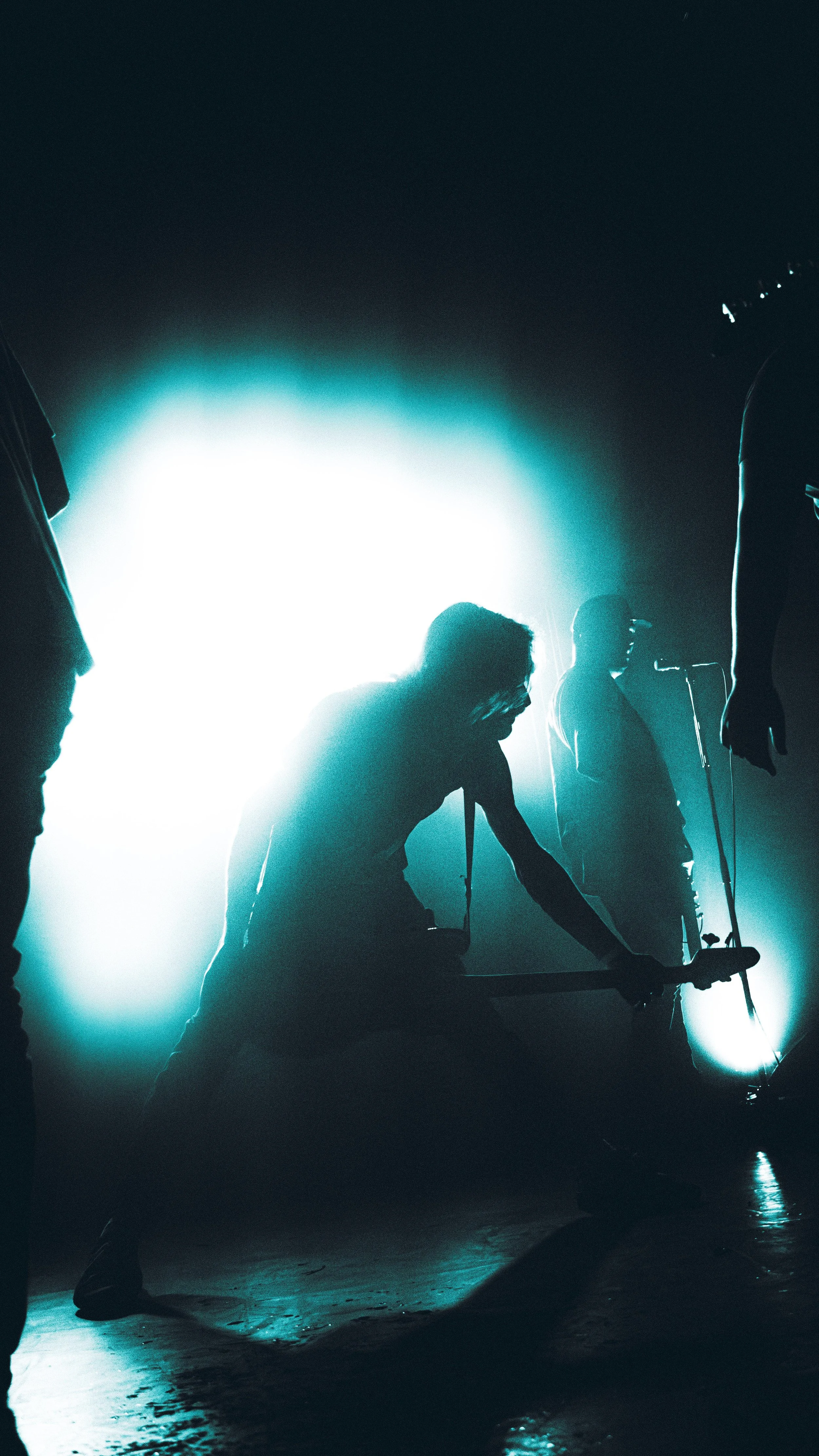 Silhouettes of several people, including one kneeling with a microphone stand, backlit by bright teal light in a dark room, creating a dramatic scene.