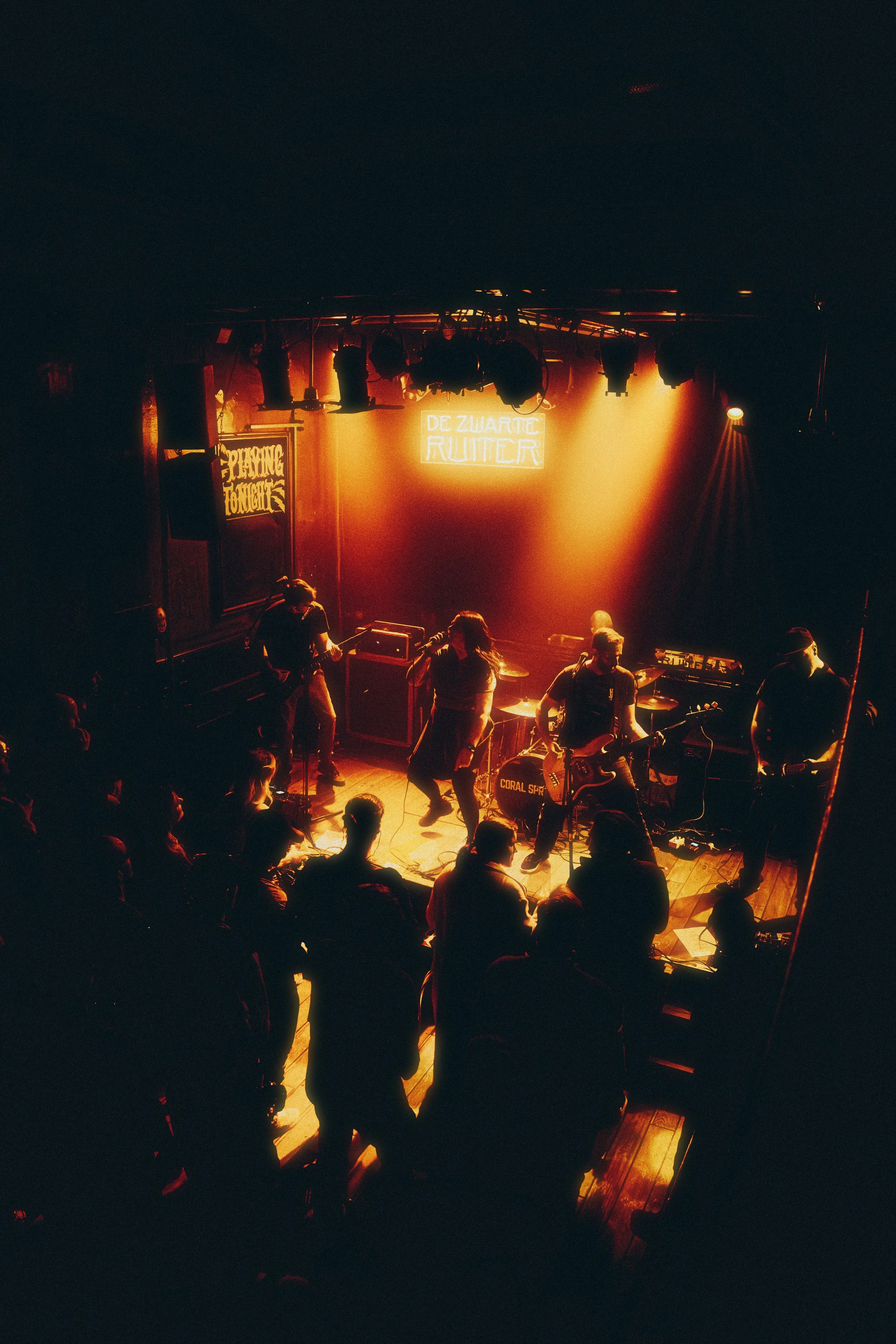 A live band performs on stage in a dimly lit venue with orange and yellow lighting, with an audience standing in front of them.