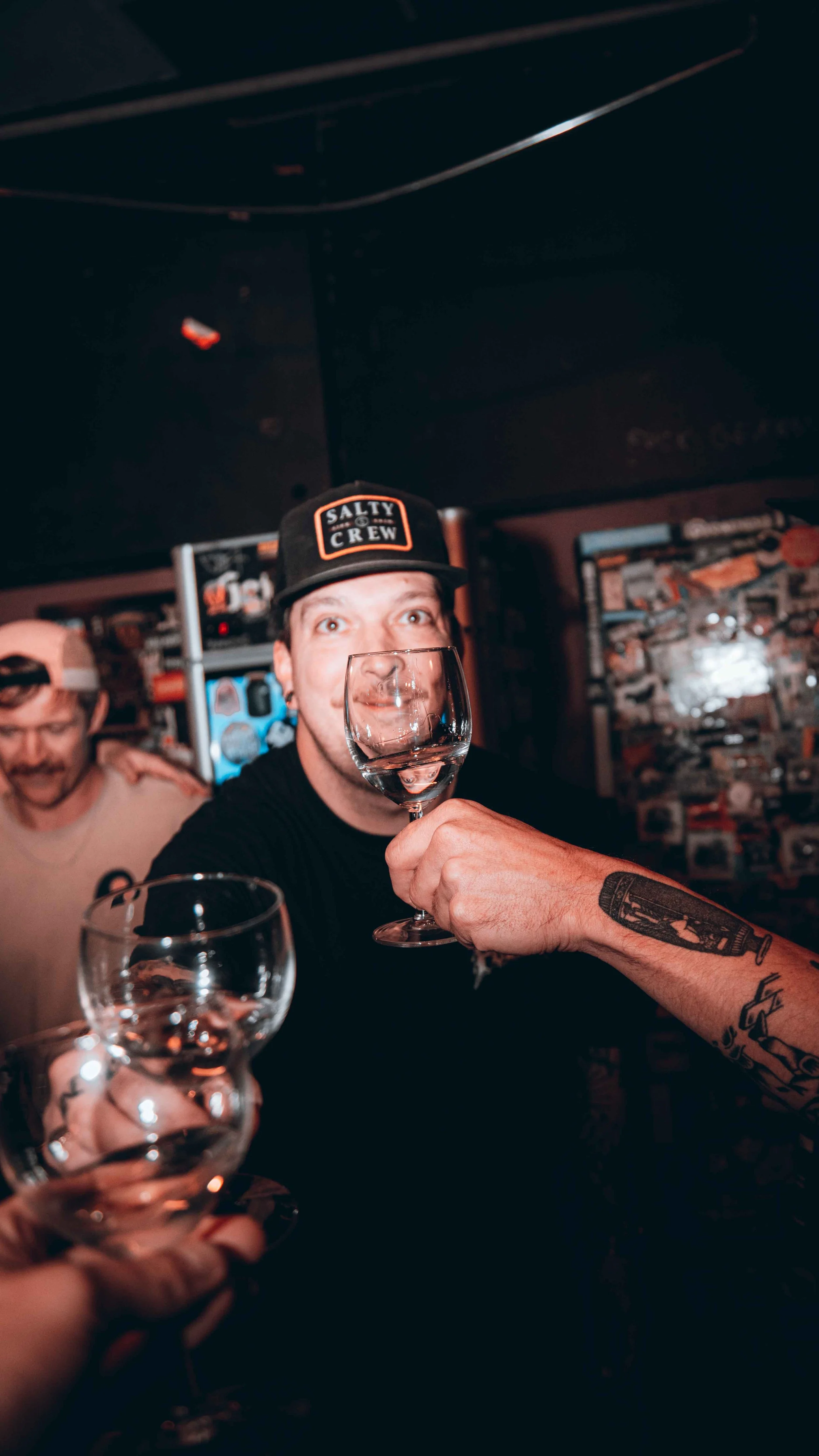 A man wearing a black cap with 'Salty Crew' is holding a wine glass towards the camera at a festive gathering. Another person in the background is smiling, and another hand holding a similar glass is visible in the foreground. The setting appears to 