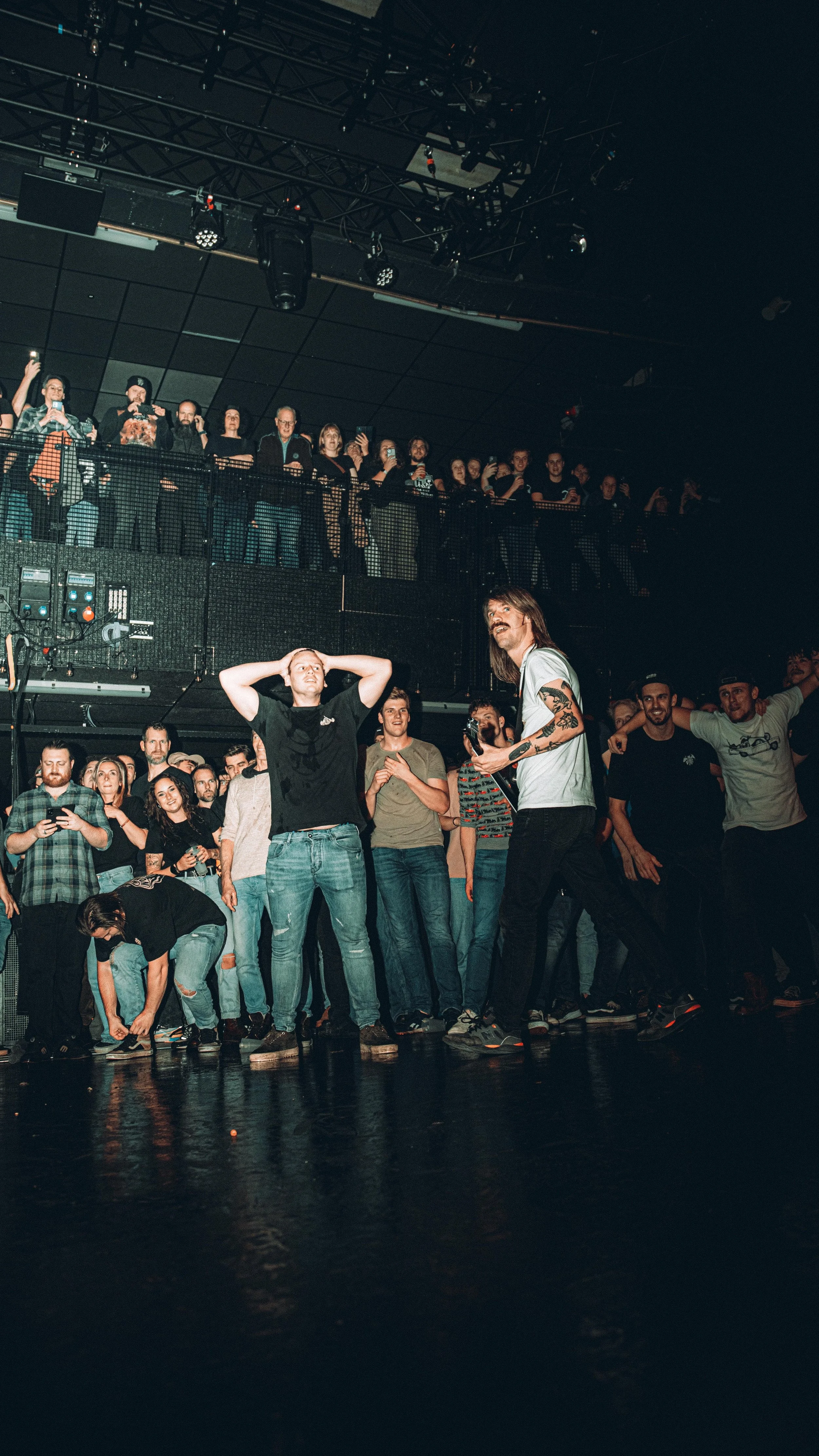 A crowd of people at a concert or live event, with some on the ground level and others on an upper balcony, watching and taking photos. Two men are in the foreground, one with his hands behind his head and the other holding a guitar, surrounded by ot