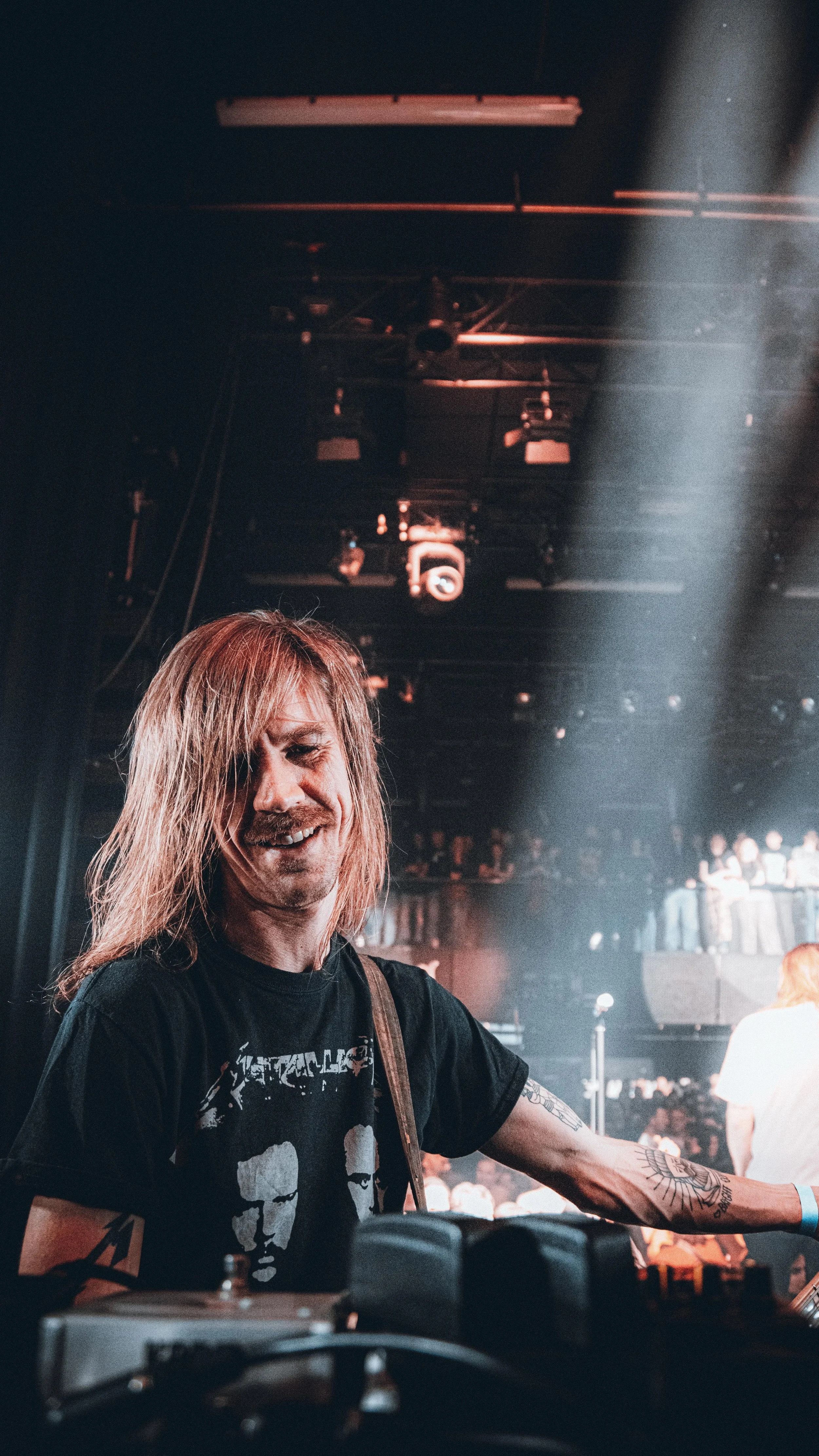 A male musician with long blonde hair and tattoos on his arm performing on stage at a concert, with a dark background and audience in the distance.