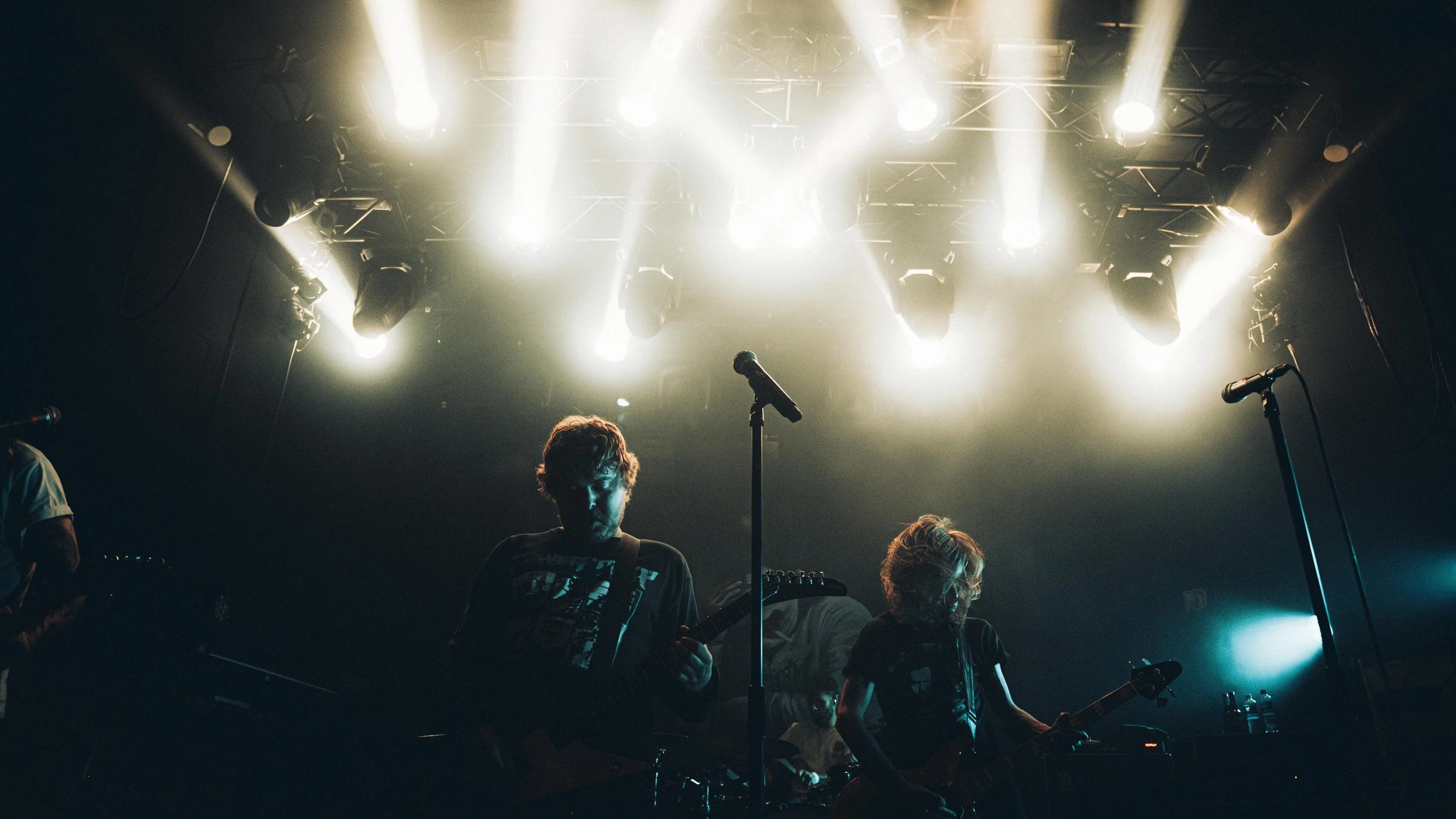 Musicians performing on stage with bright overhead lights, two guitarists in the foreground, silhouette visible in dark lighting.