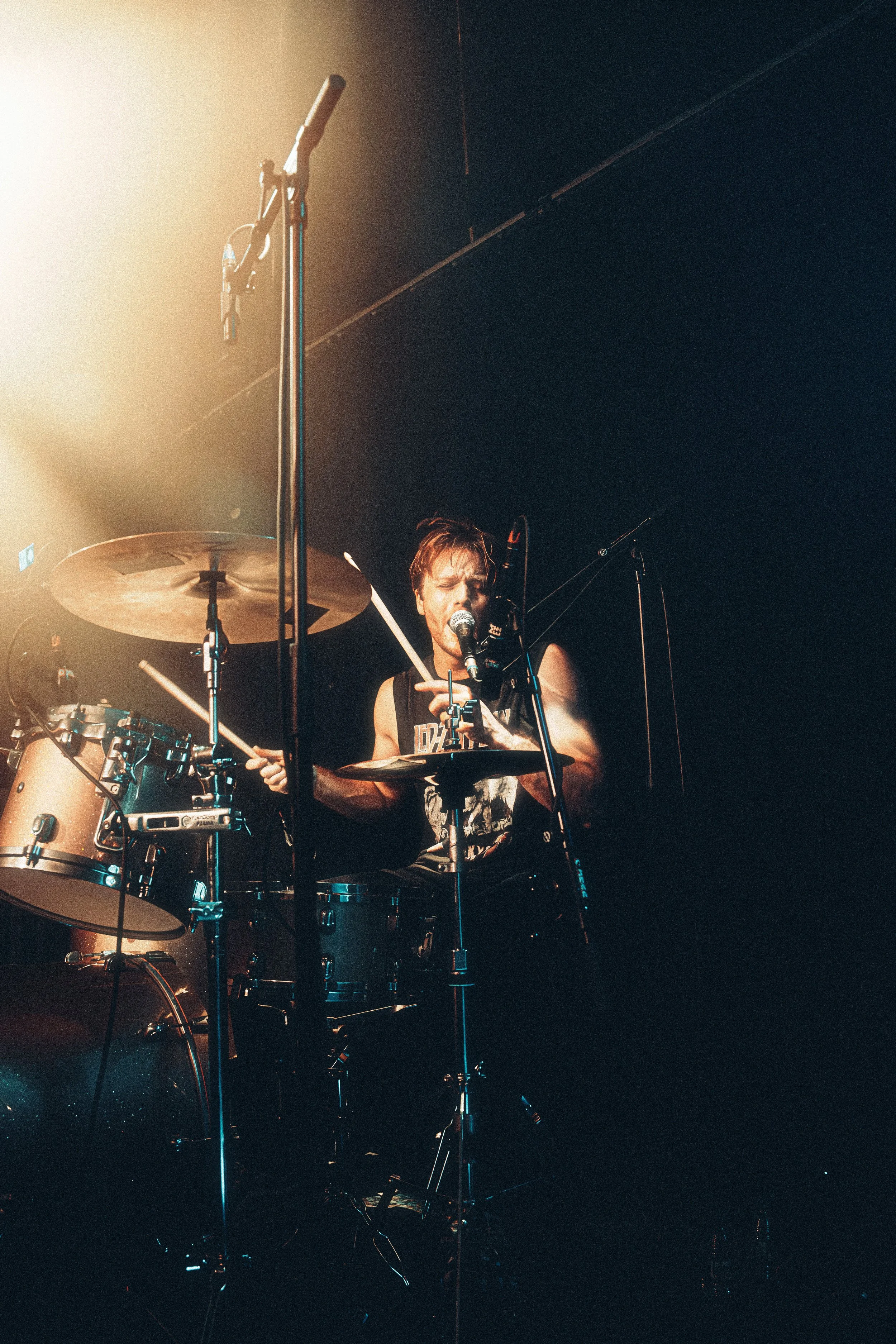 A drummer playing drums on stage, singing into a microphone, with stage lights illuminating him.