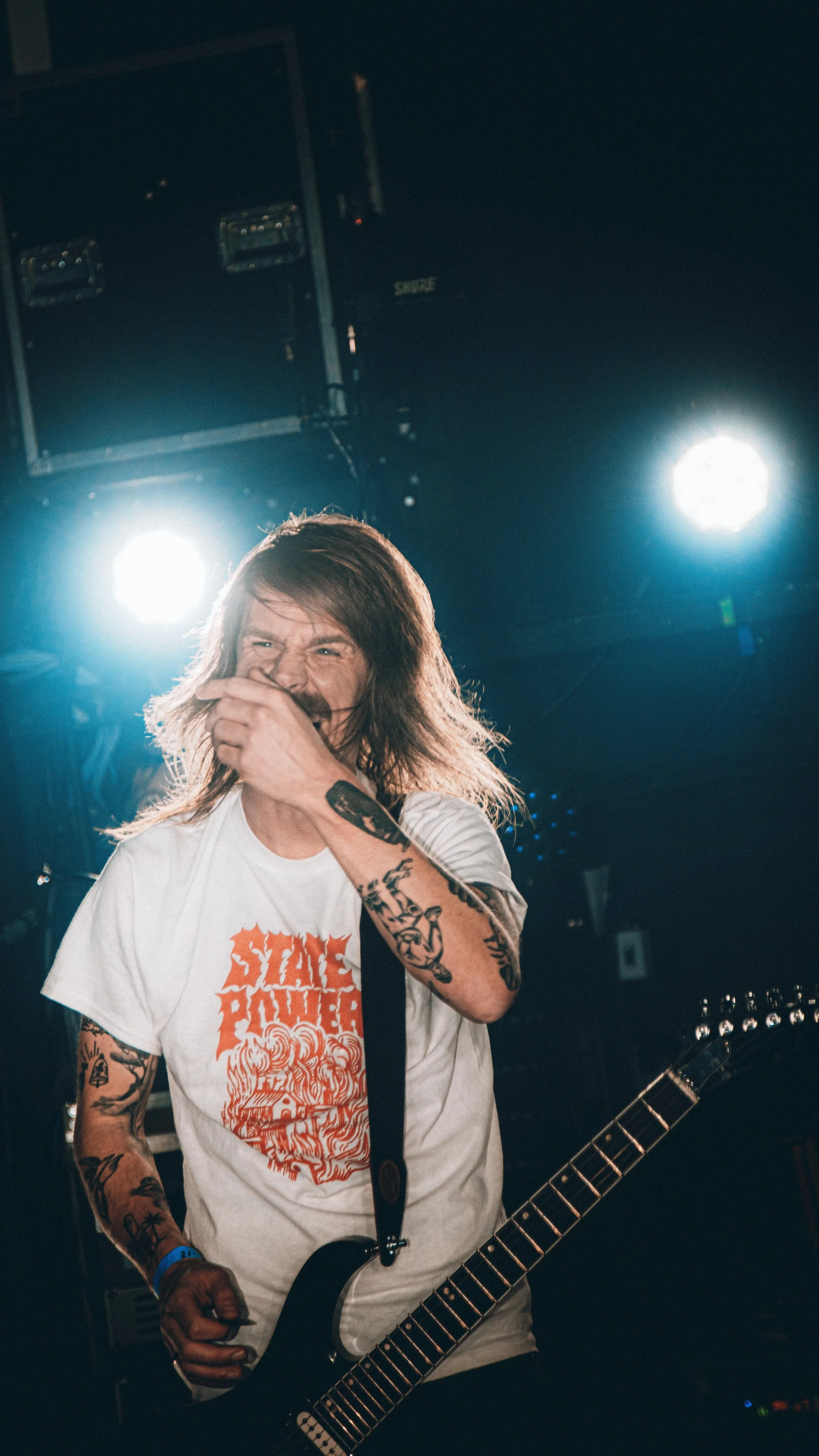 A male guitarist with long hair, tattoos, and a white shirt with orange text, laughing and covering his mouth while playing a black electric guitar on stage, with bright stage lights behind him.