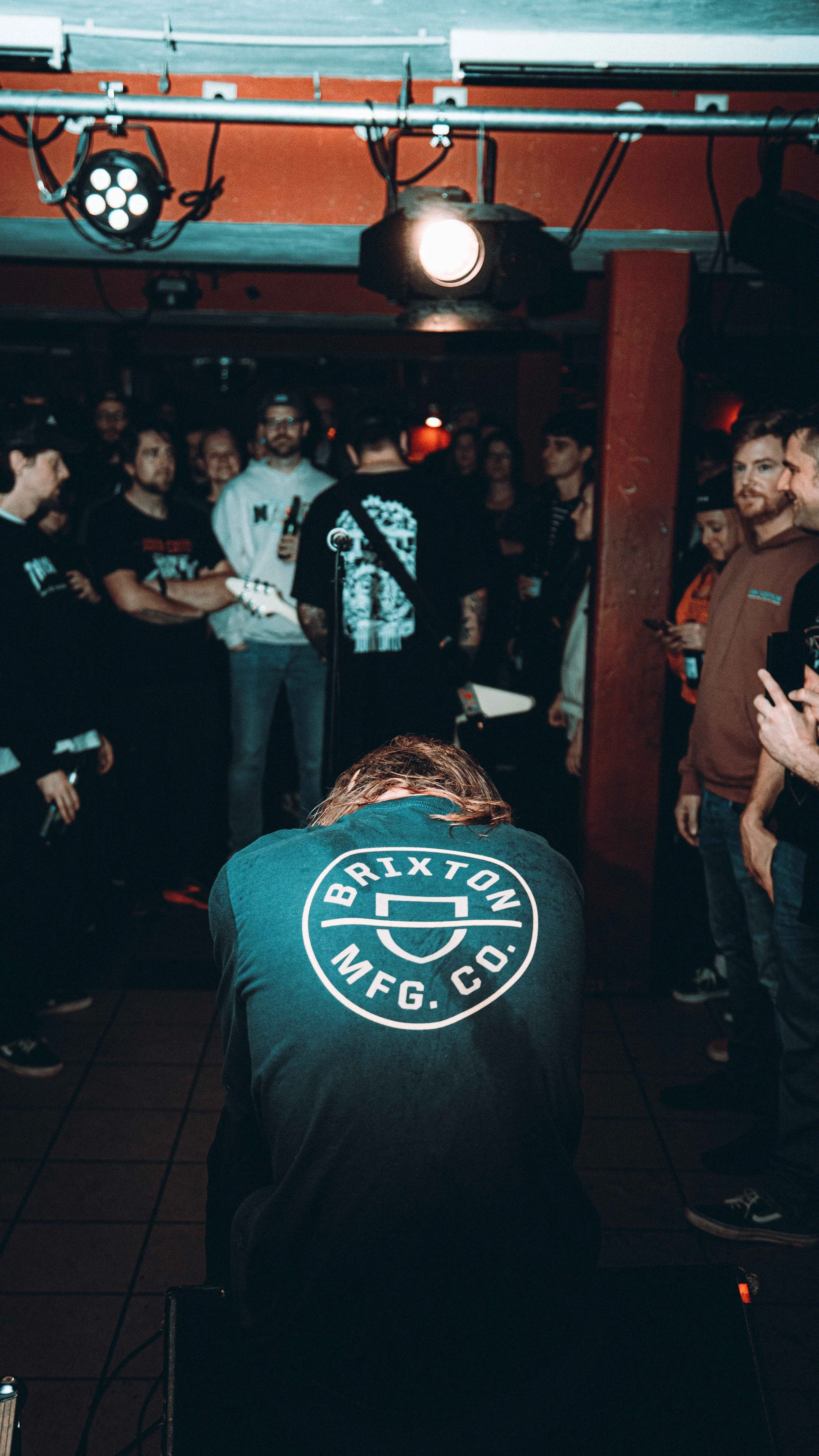 A person with long hair kneeling with their head down, wearing a black shirt with the logo 'BRIXTON MFG. CO.' on the back, in a concert or event setting with a crowd of people standing and watching.