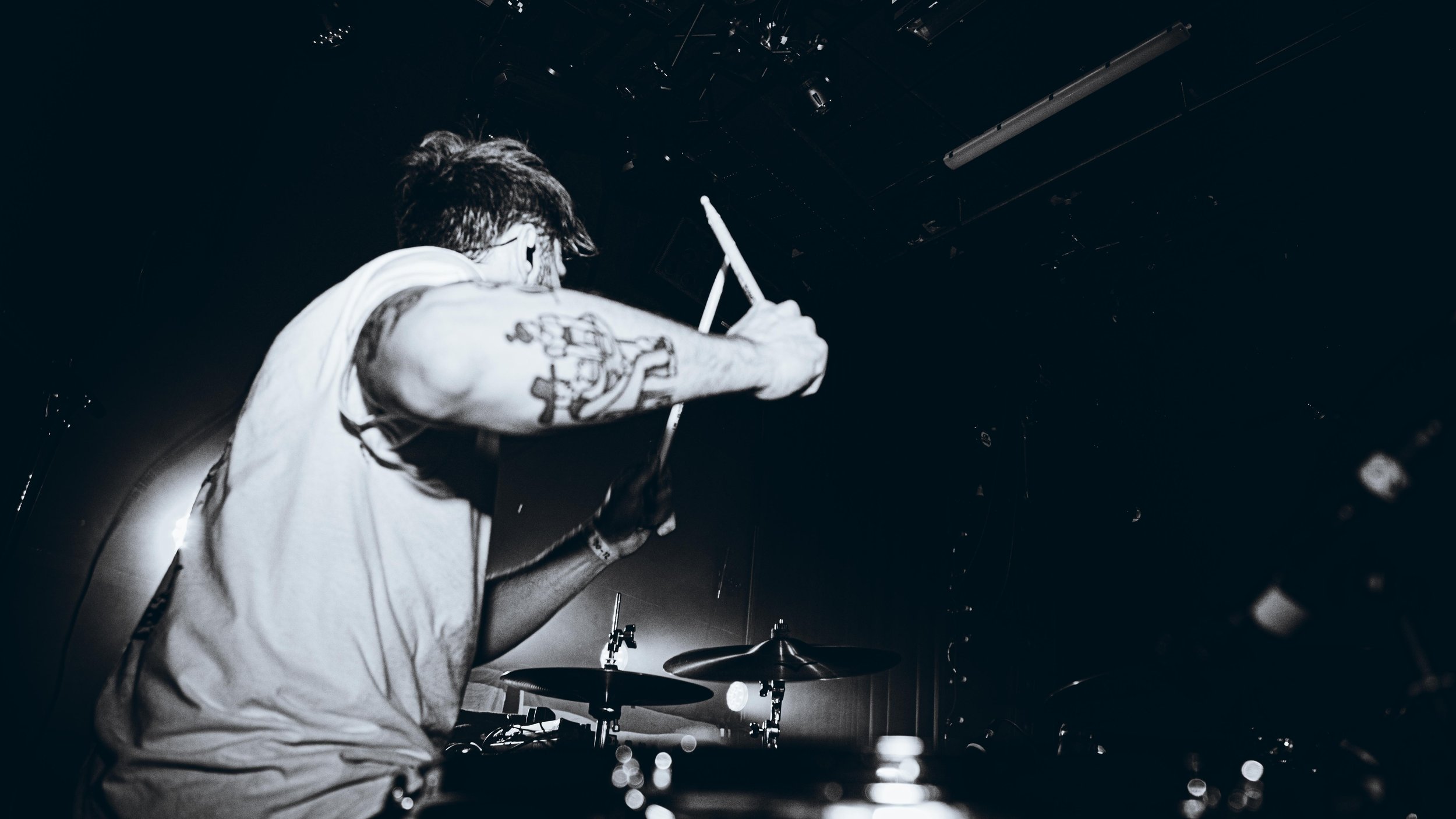 A man with tattoos on his arm playing drums on stage, viewed from behind, in black and white.
