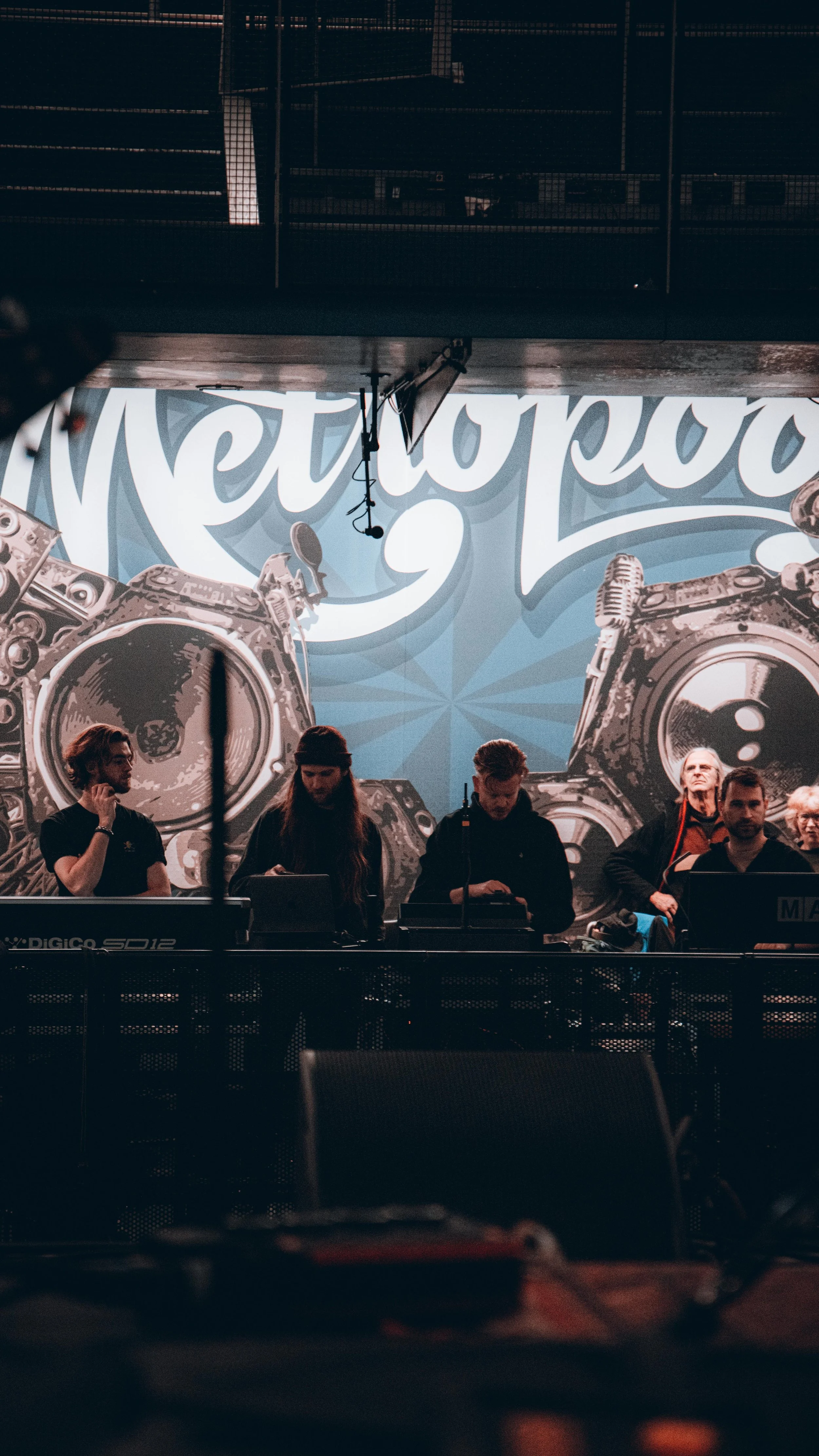 A group of people standing behind DJ equipment on a stage, with a large mural of speakers and the word "Metropole" in the background.