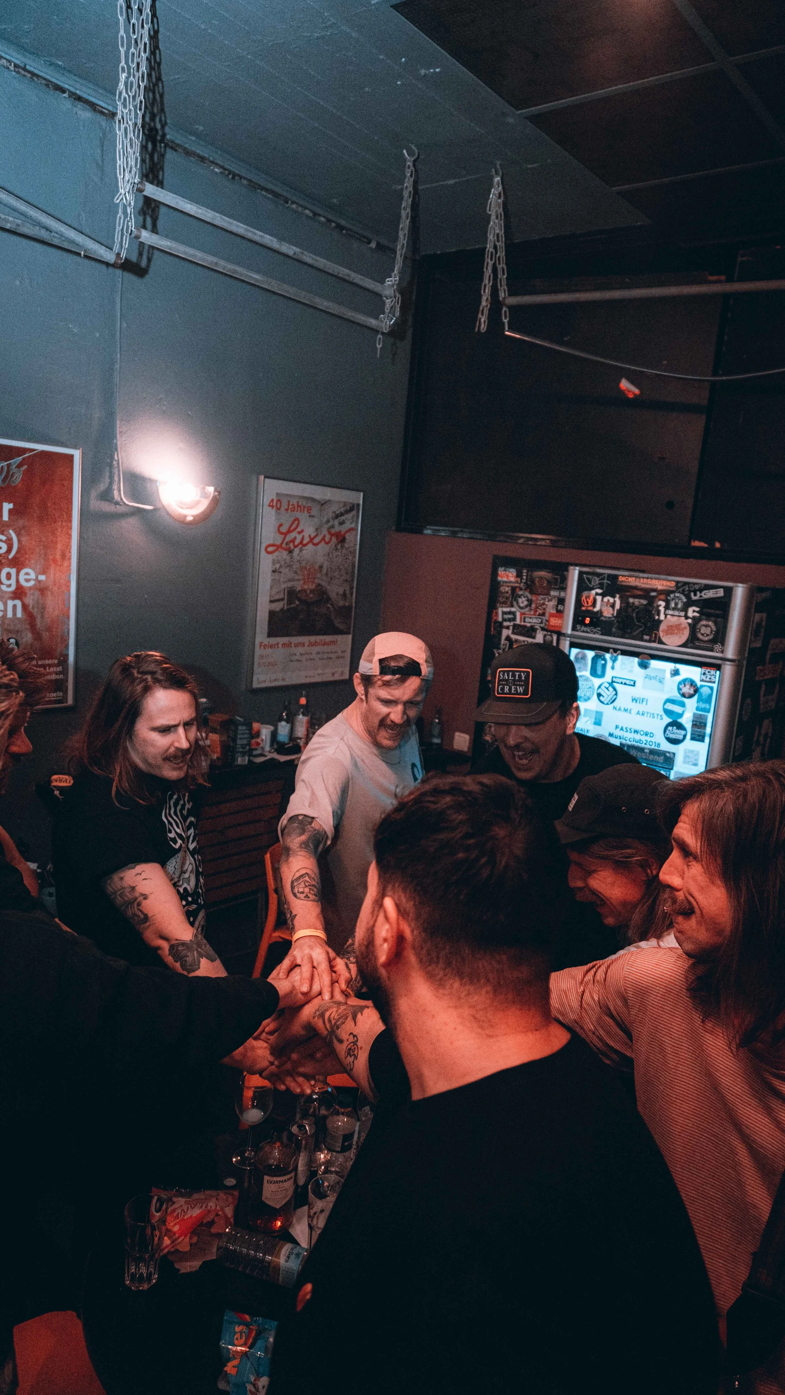 A group of friends cheer and hold hands in a circle at a bar, with drinks on the table, celebrating together.