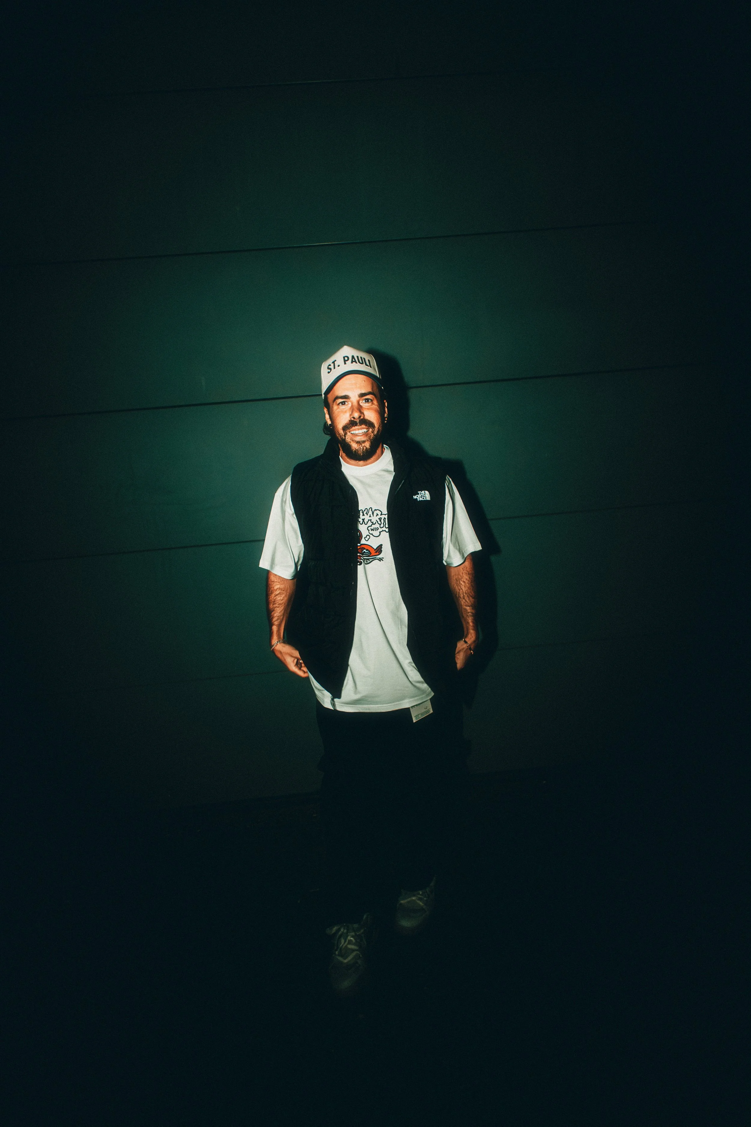 A man standing in front of a dark wall, wearing a hat with 'St. Pauli' written on it, a white T-shirt, a black vest, and dark pants.