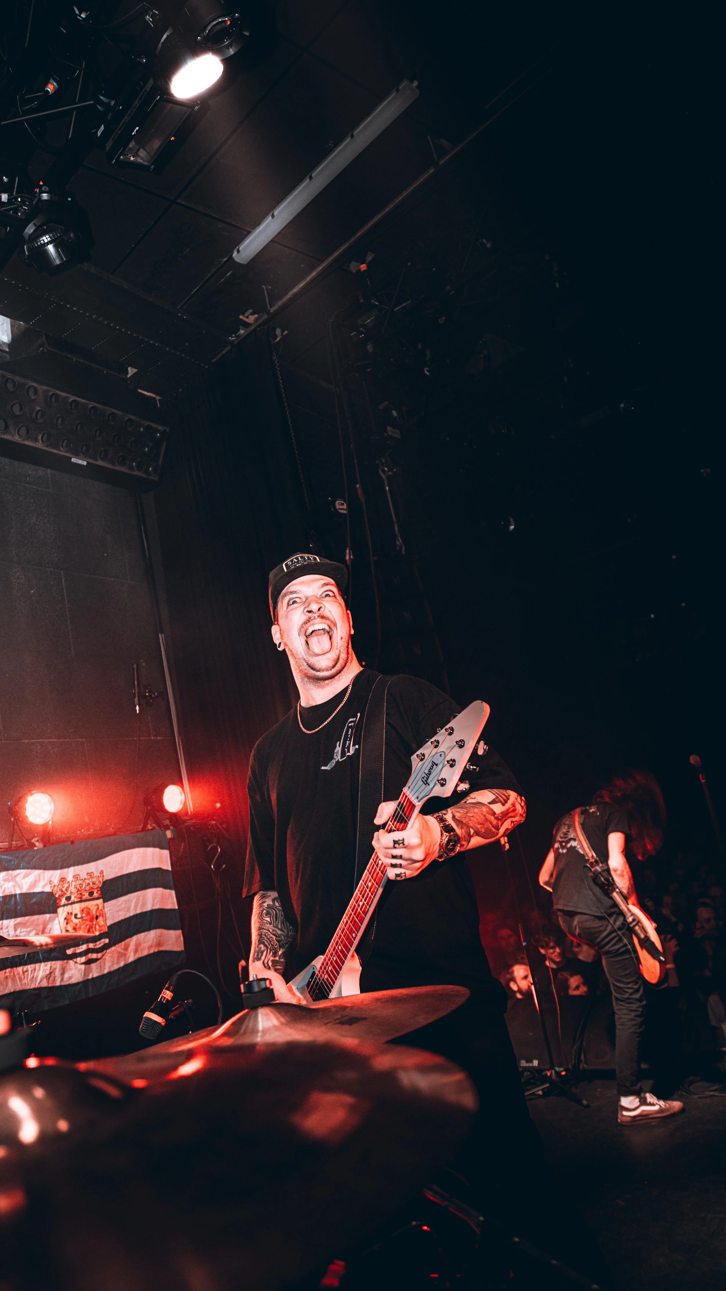 Musician playing guitar on stage during a concert with red lighting, a drummer's cymbal in the foreground, and a bandmate in the background.