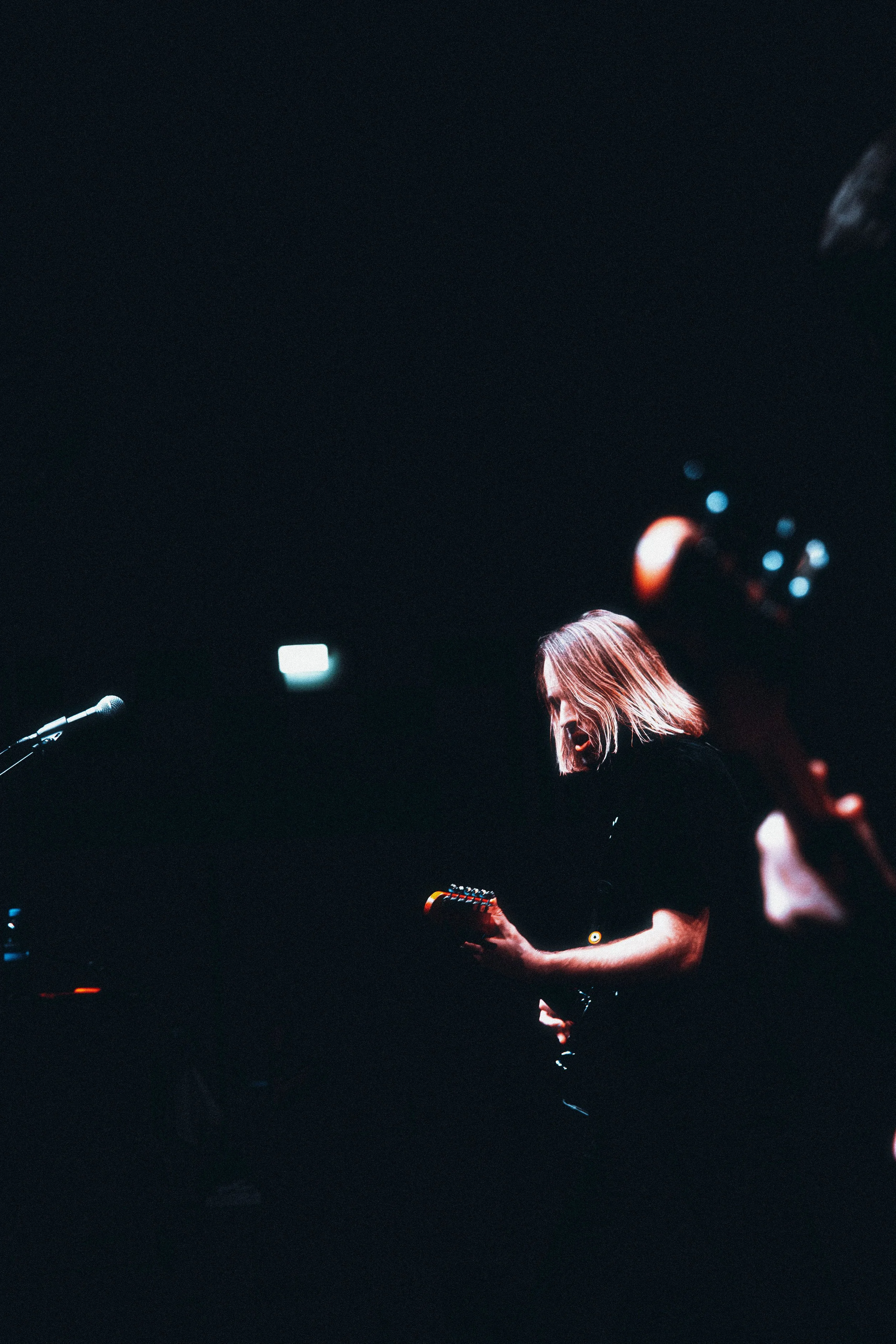 A woman playing an electric guitar on stage in a dark setting, with a microphone to her left and stage lights in the background.