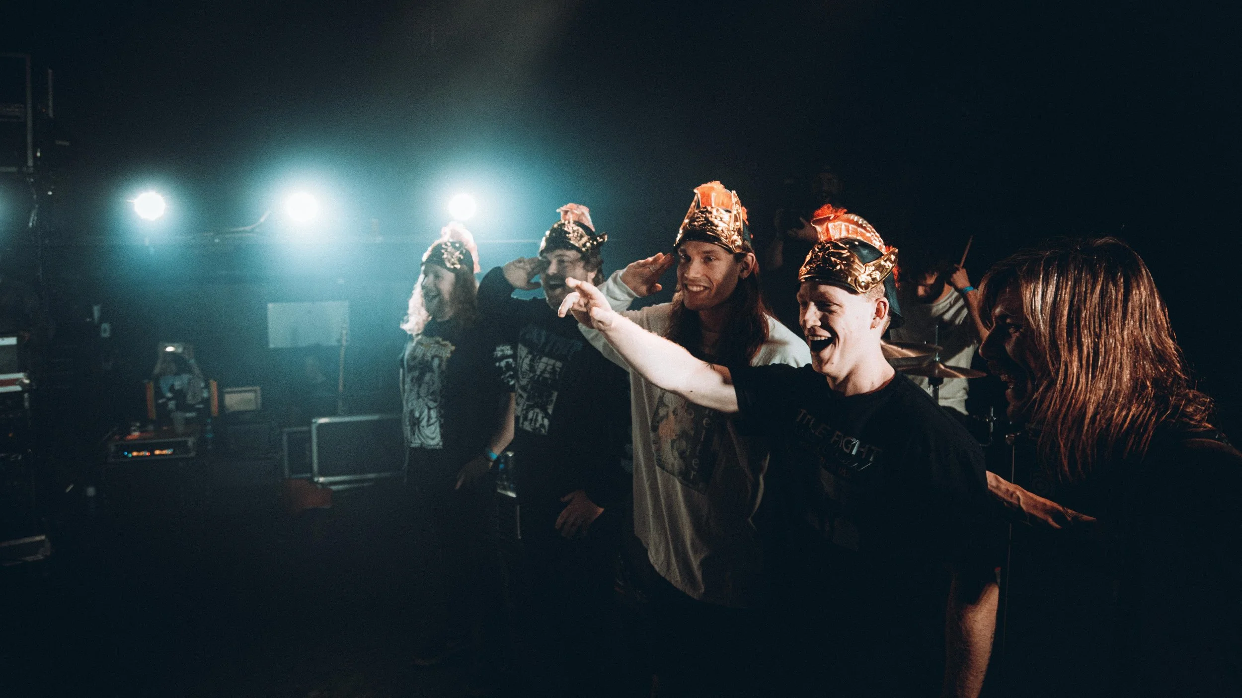 Group of young adults in party hats celebrating and smiling, backlit with stage lights in a dark indoor setting.