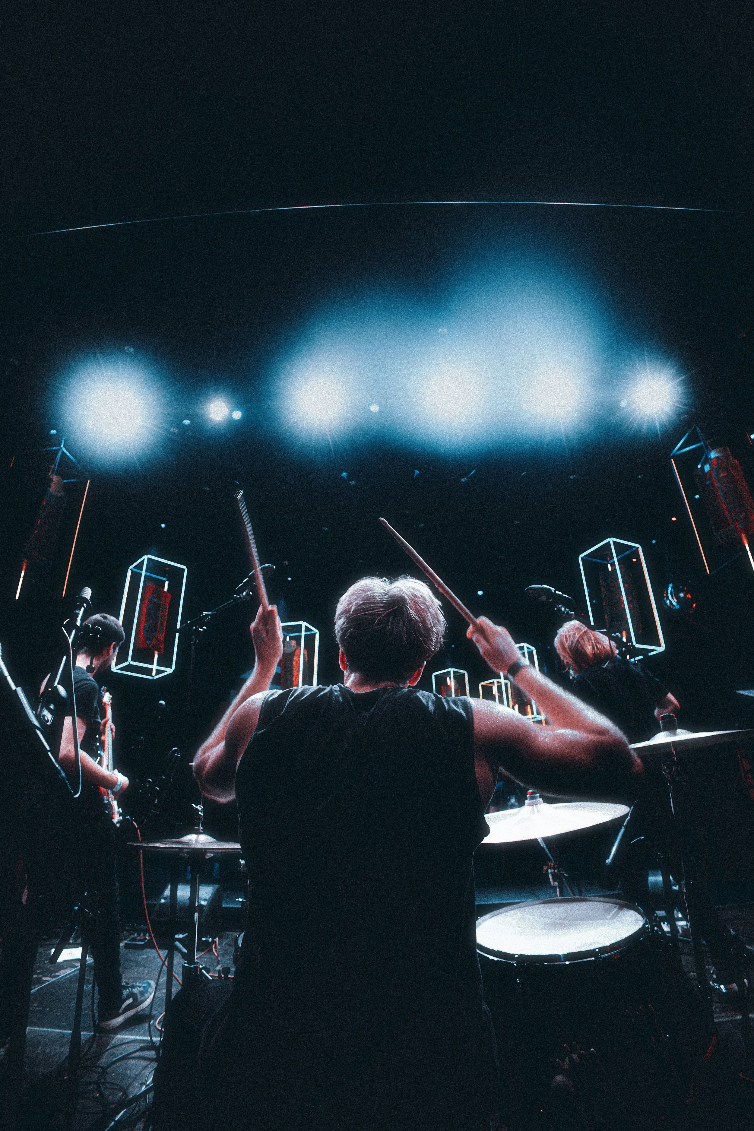 A drummer performing on stage with other band members, illuminated by bright stage lights and surrounded by electronic equipment.
