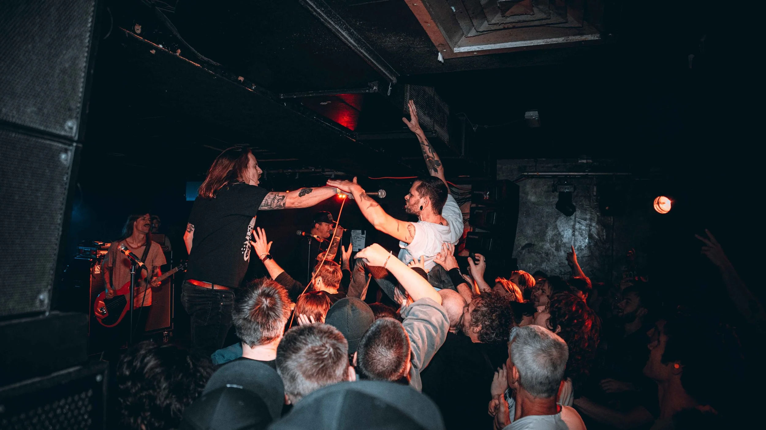 A live concert scene with a crowd of people dancing and reaching towards the stage, where band members are performing. One person is crowd surfing, being held up by fans. The stage has musical equipment and dark lighting.
