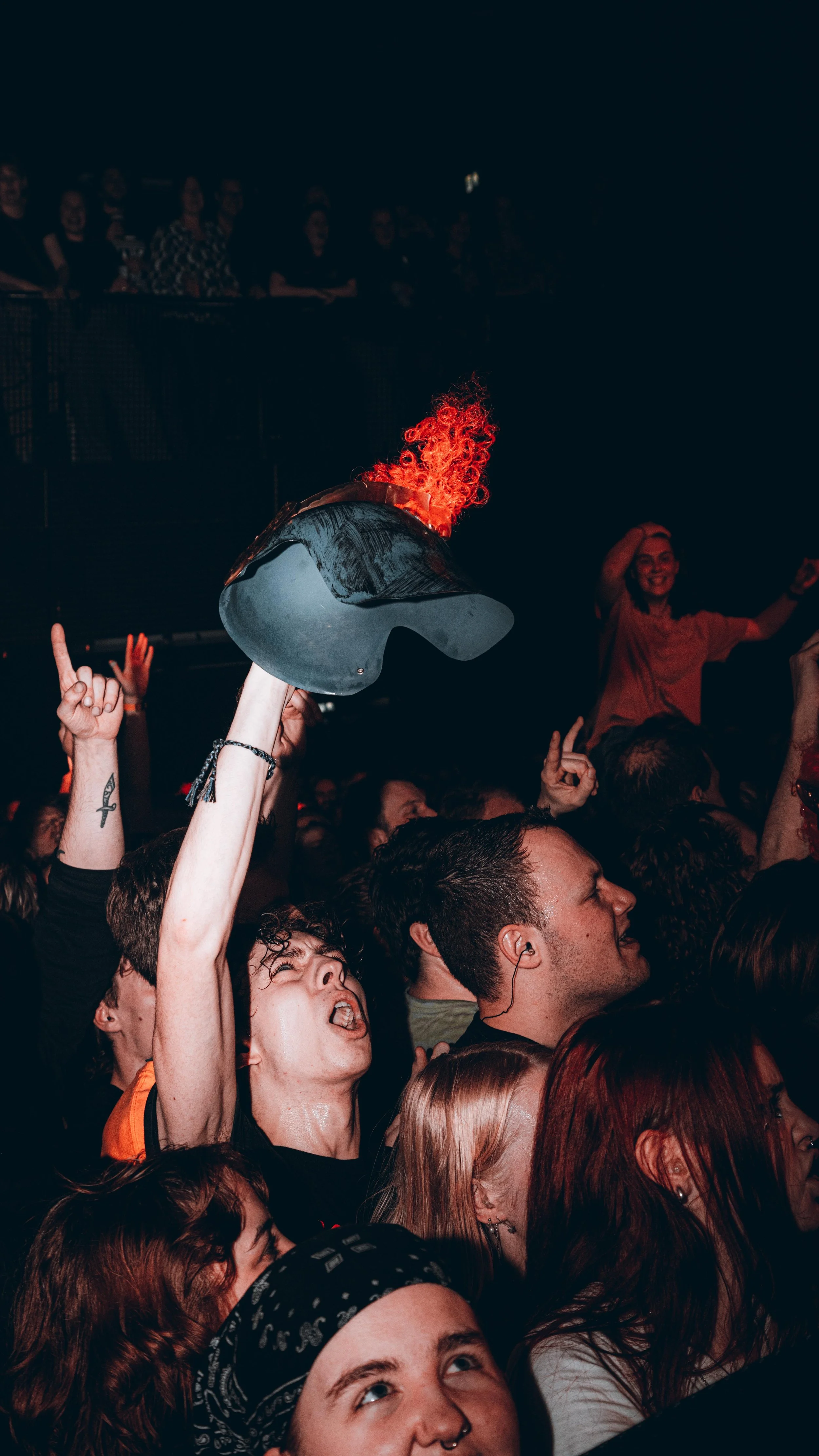Crowd of people at a concert, some with raised hands, one holding a hat with red hair attached, celebrating and enjoying live music.
