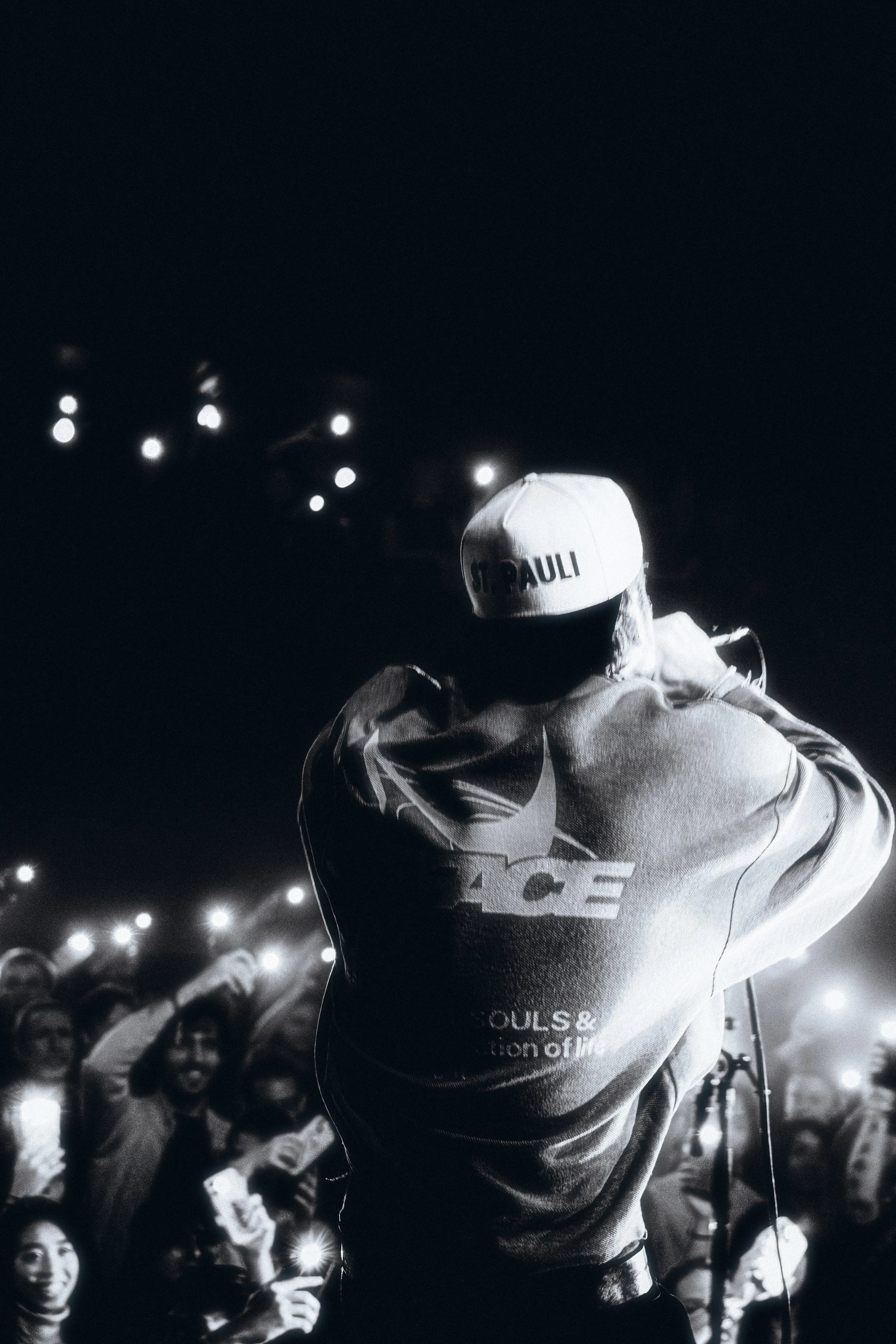 A person wearing a white cap with 'ST. PAULI' written on it and a hoodie with a large logo, facing an audience at night in a dimly lit outdoor setting.