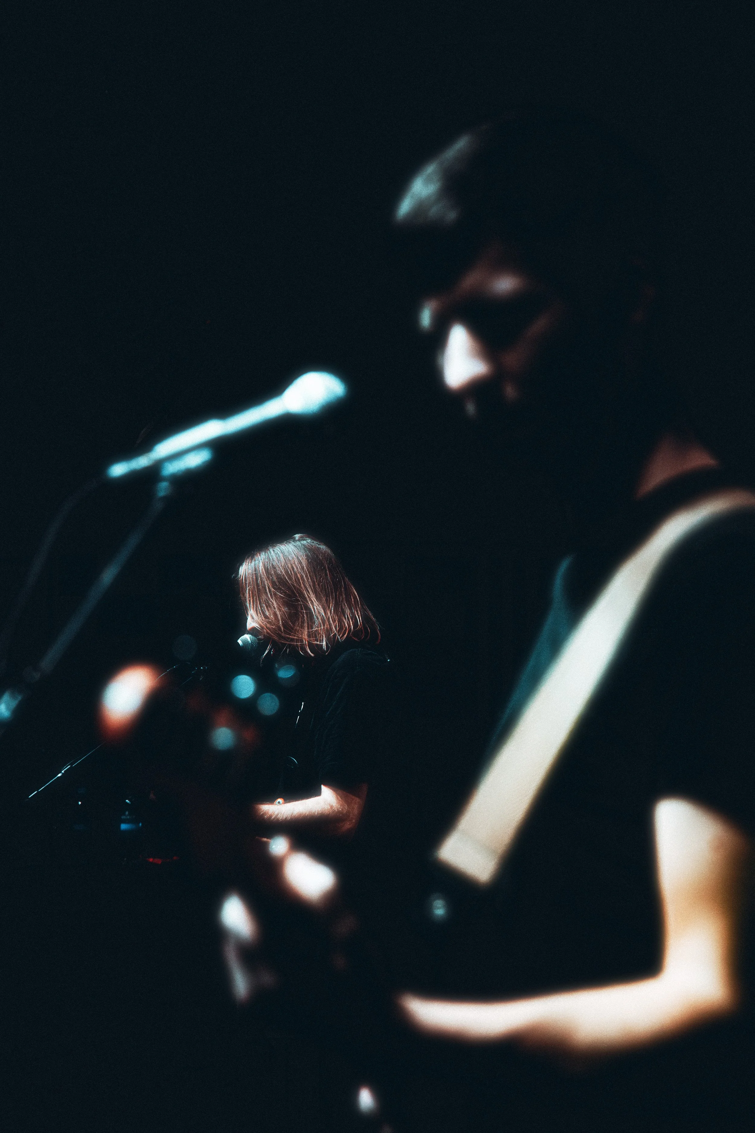 Two musicians performing on stage with microphones, one playing guitar in the foreground and the other singing or playing guitar in the background; dark lighting enhances the scene.