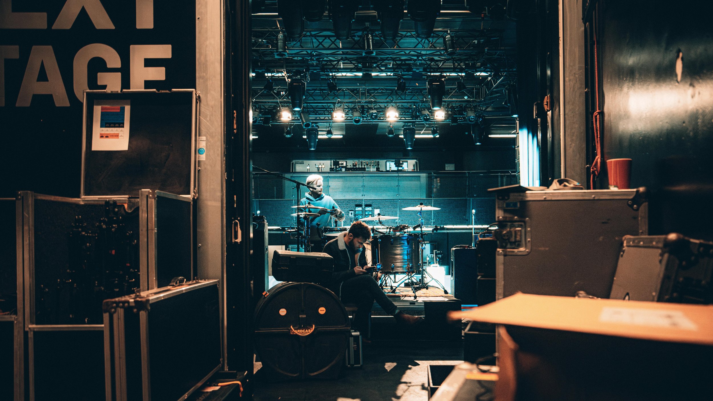 View into a small stage area with two musicians preparing, one sitting with a laptop and the other behind drums, surrounded by equipment and stage lights.