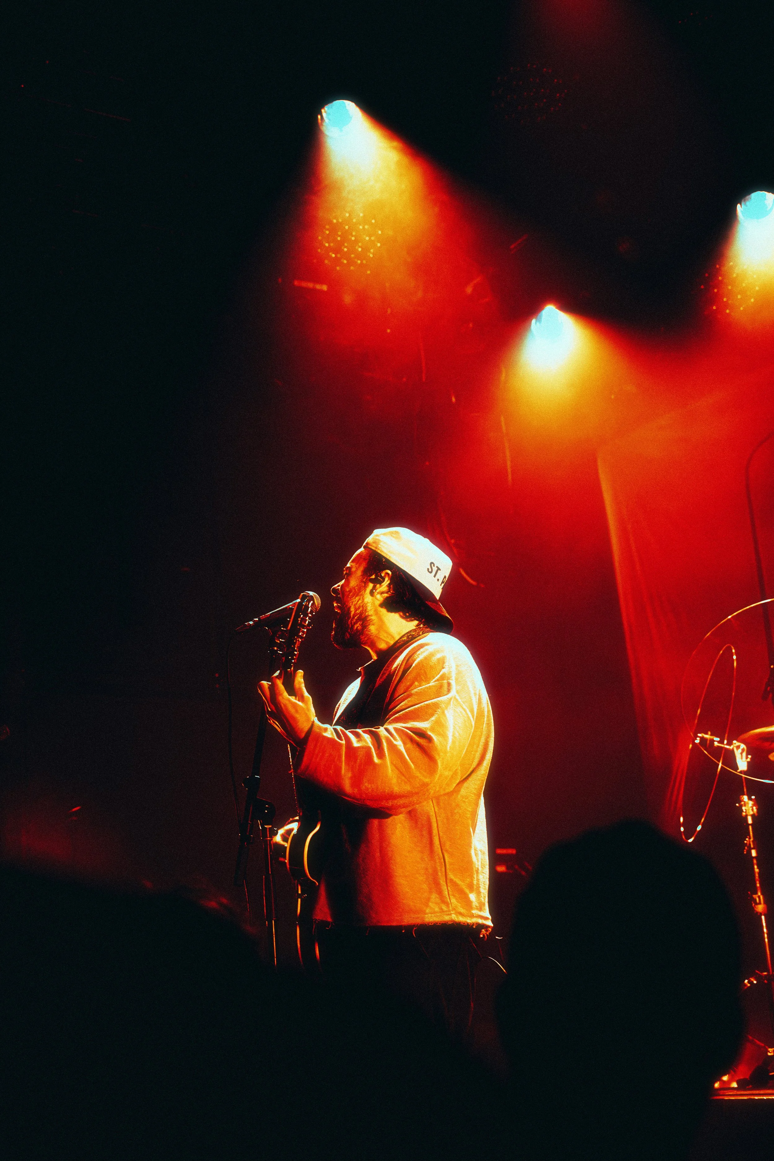 A musician performing on stage under orange and blue stage lighting, wearing a backward cap and jacket, singing into a microphone while playing a guitar.