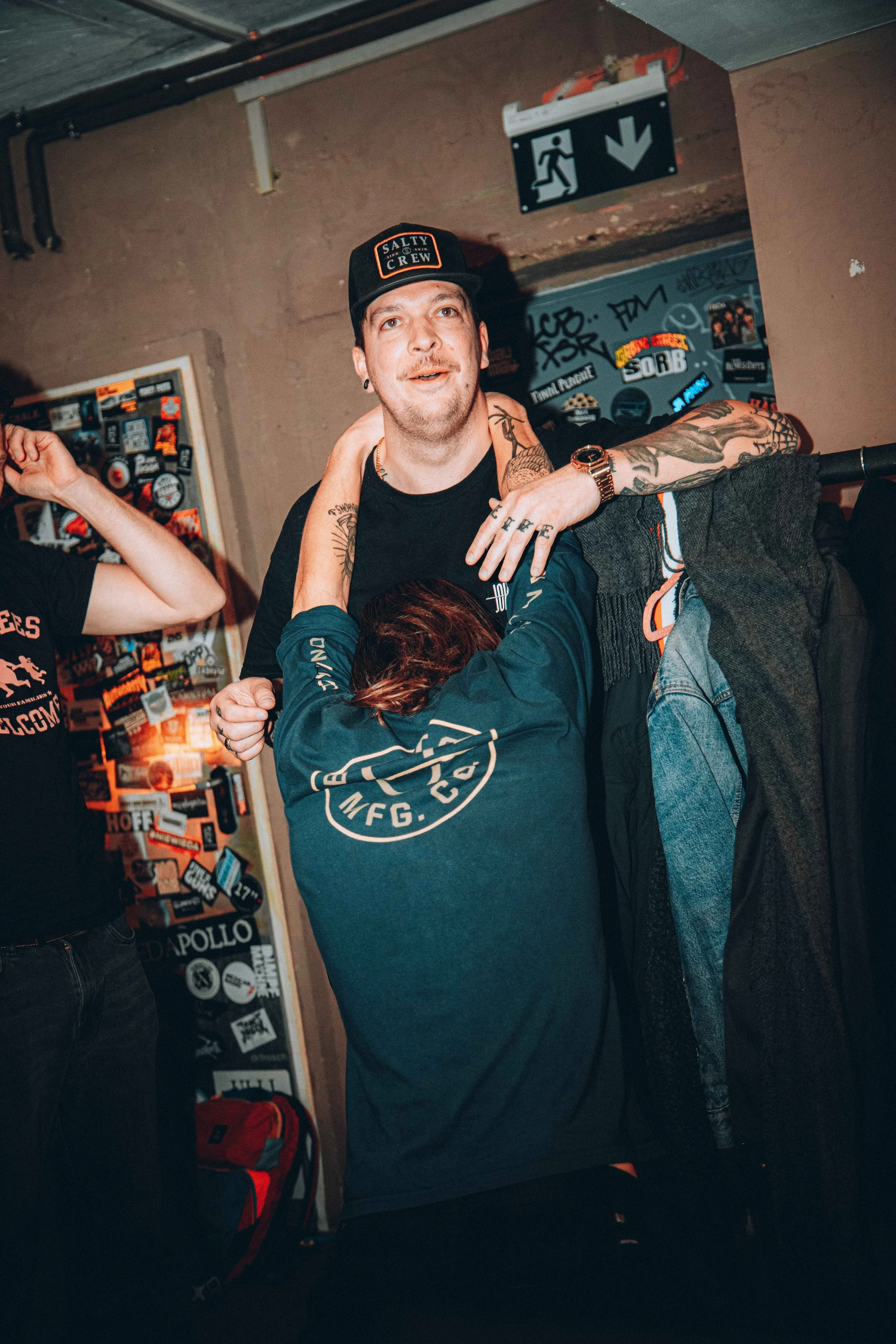 A man with tattoos and a cap, standing with a woman who has her arms around his neck, in a room decorated with stickers and patches. The man is smiling and looking at the camera, while the woman has her head down.