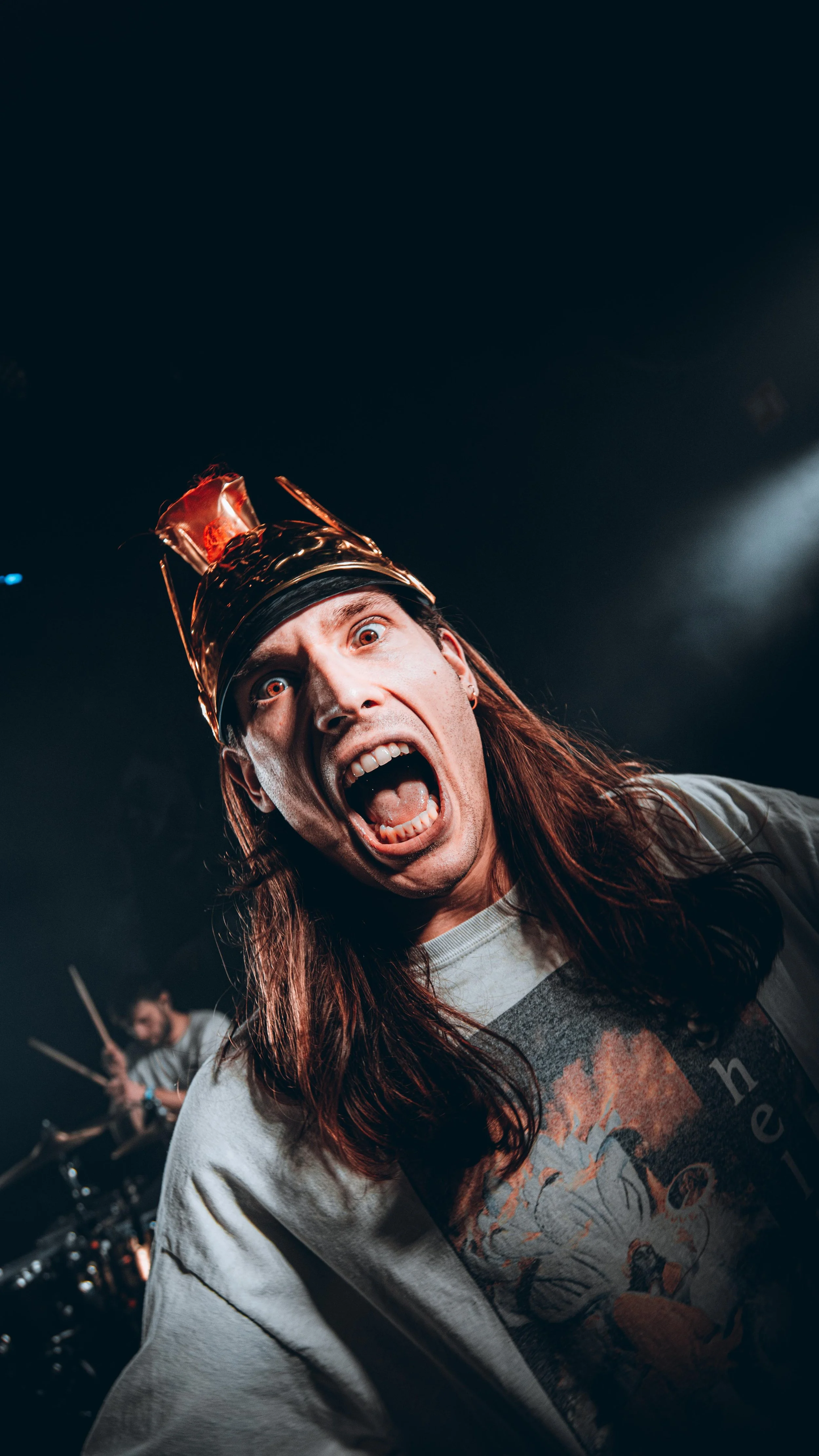 A man with long hair wearing a metallic crown, shouting with his mouth wide open on stage, with a drummer playing in the background.