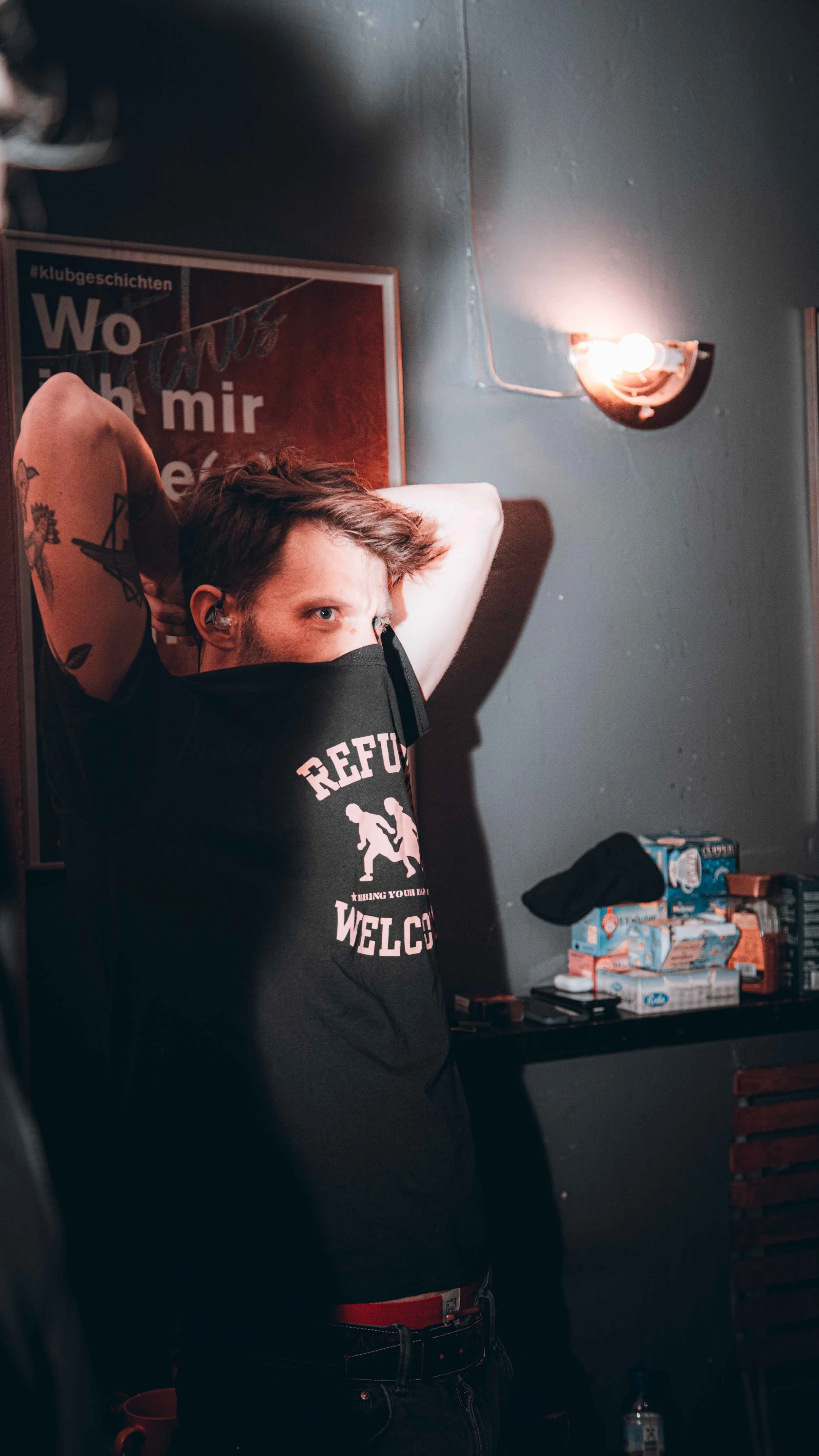 Young man with tattoos on arms wearing a black T-shirt with logo, standing indoors with hands behind head, near wall light, and shelves with snacks and a water bottle.