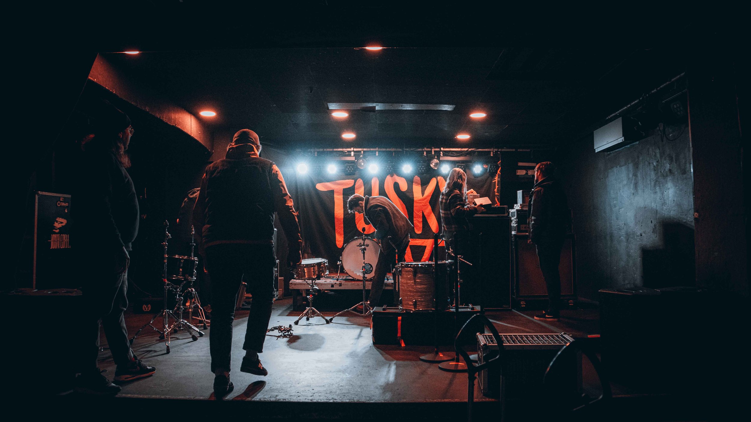 People setting up musical instruments on a dimly lit stage with a large banner reading 'TUSK' in the background.