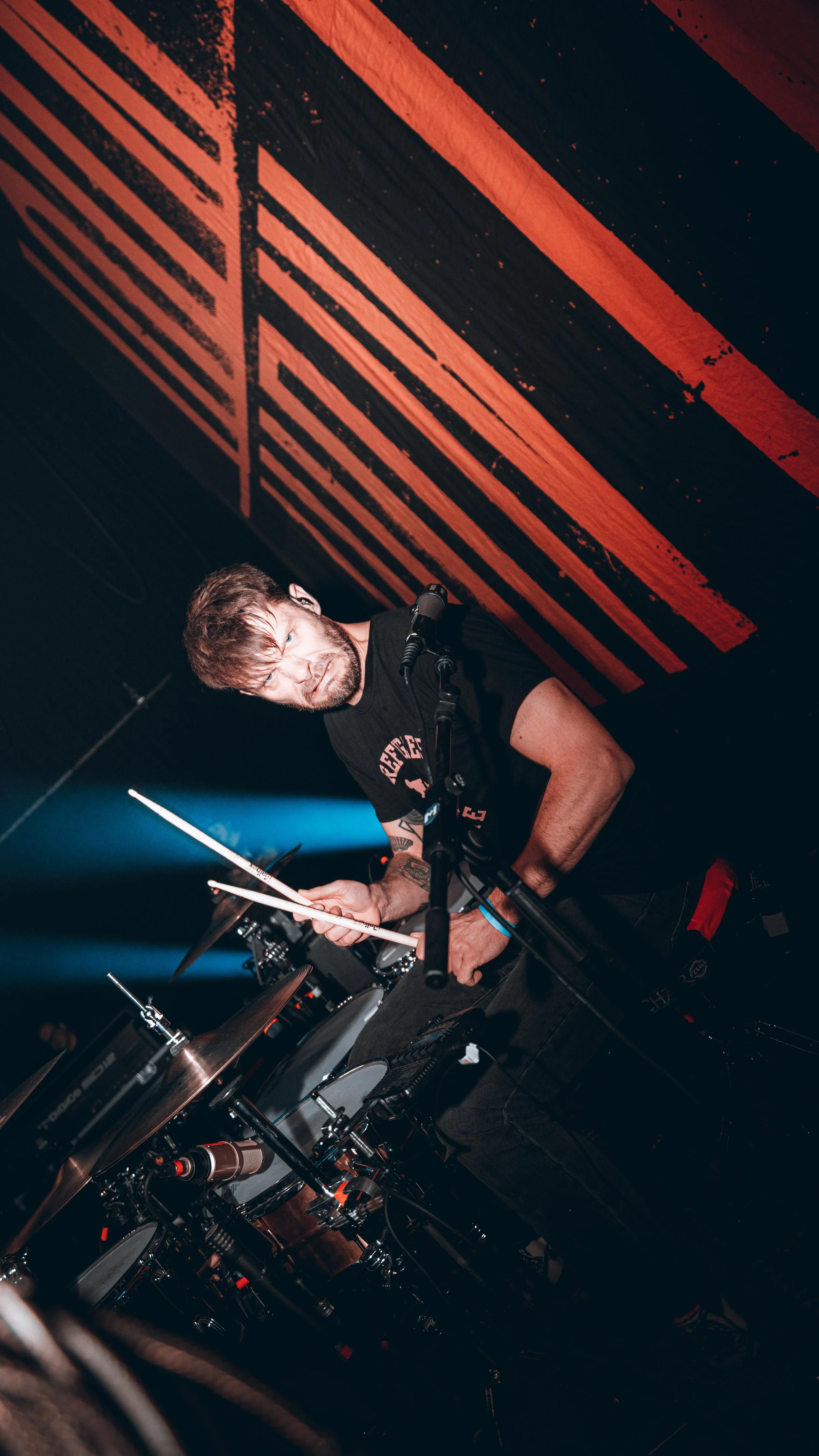 A male drummer playing a drum set on stage during a concert, with a black backdrop that has red graphics.