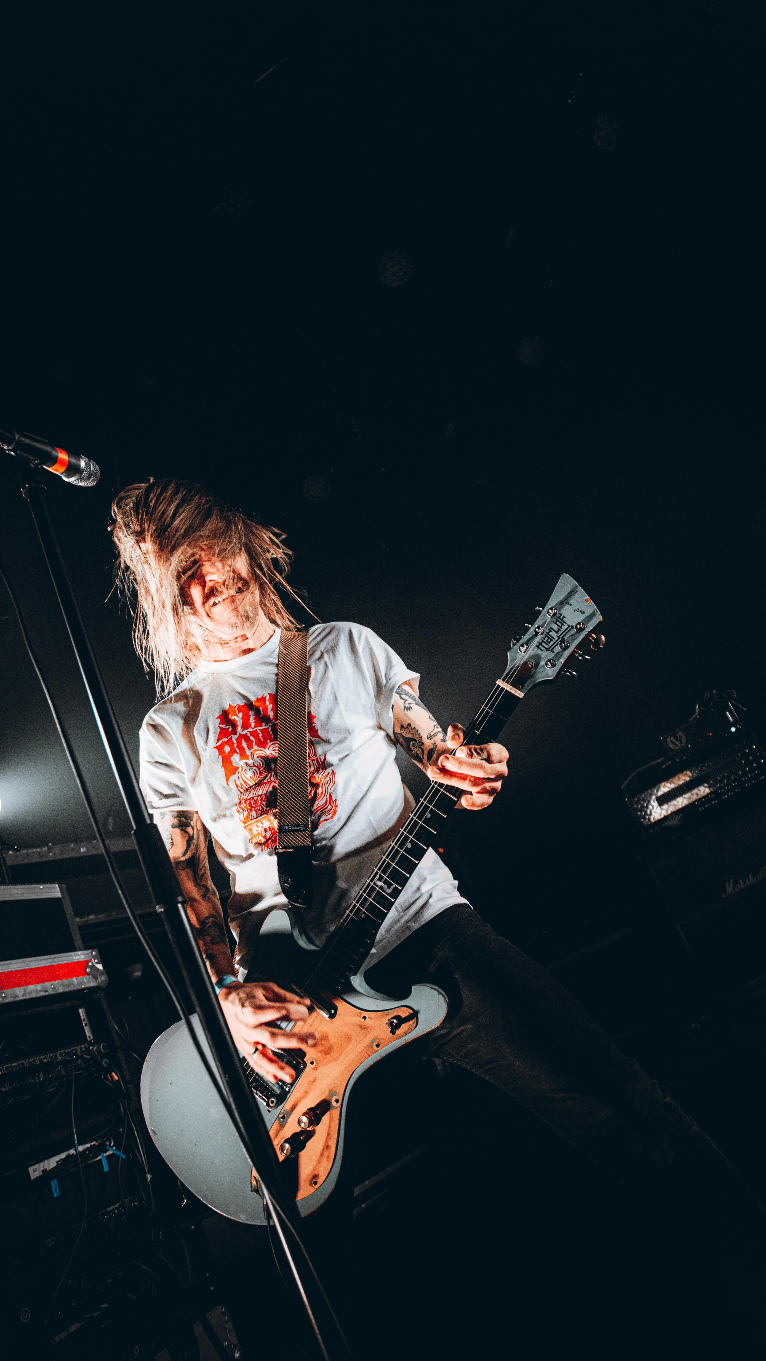 A musician performing on stage with long hair, wearing a white t-shirt with a red graphic, playing an electric guitar.